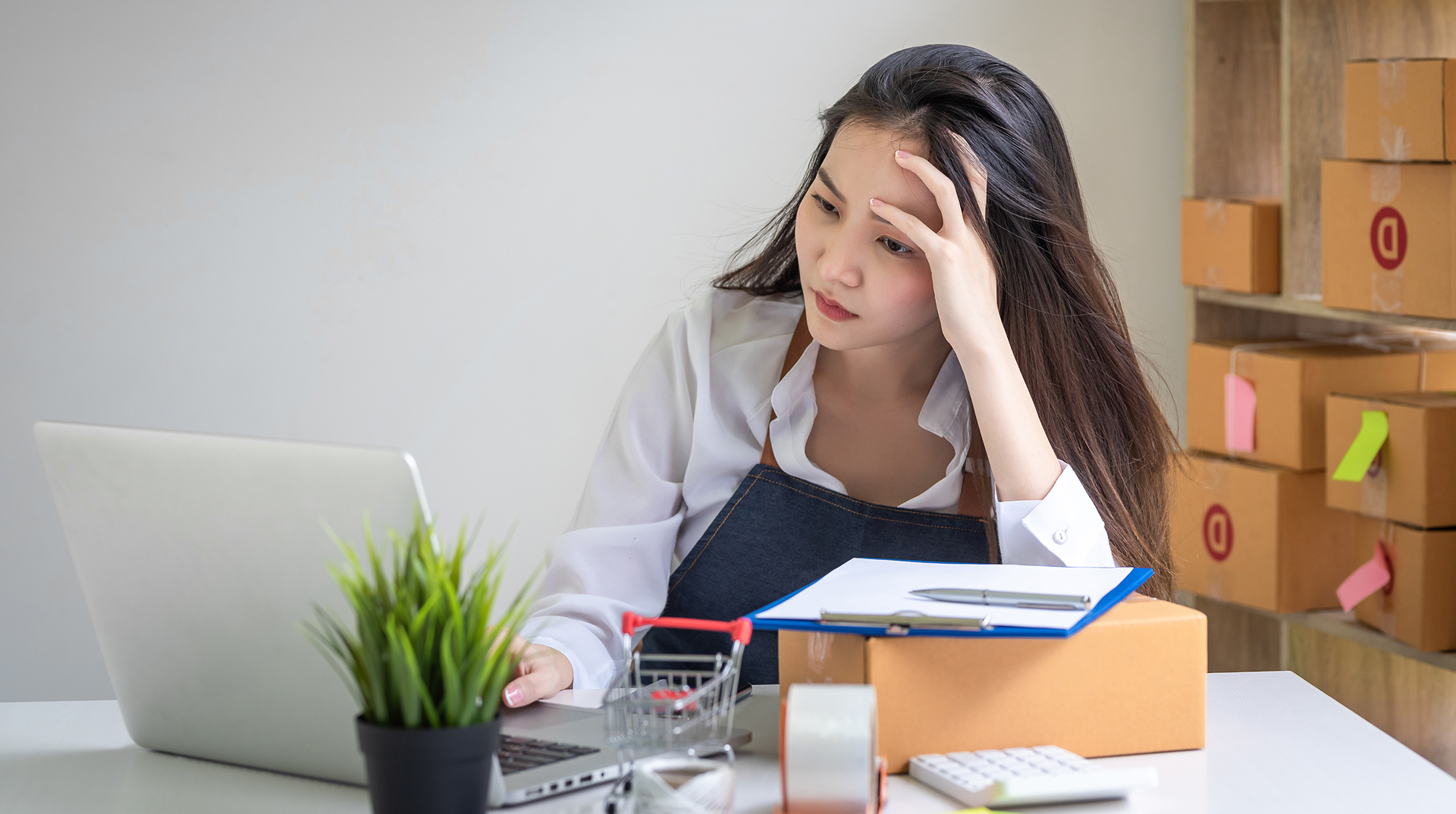 Young woman in apron working on a laptop at a desk surrounded by shipping boxes, looking concerned.