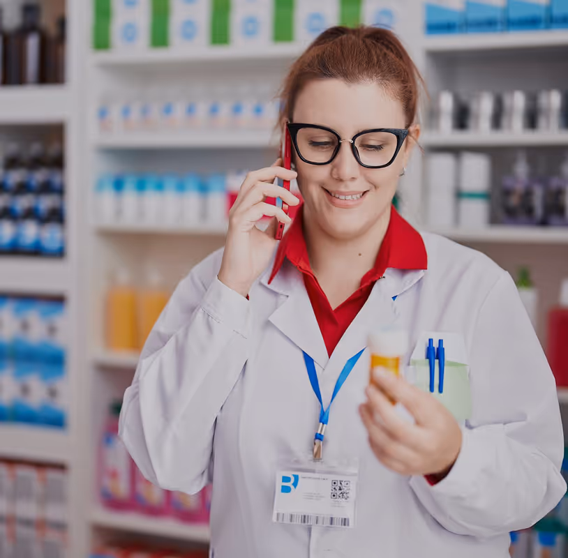 Pharmacist wearing glasses and white coat talking on phone while holding a prescription bottle in a pharmacy.