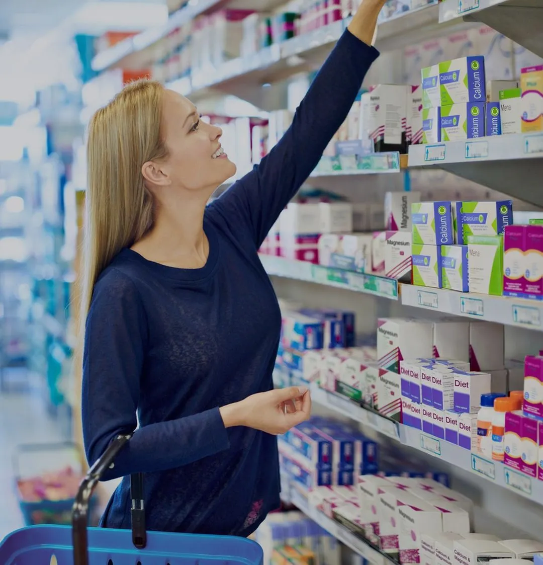 Woman in a navy blue shirt reaching for a product on a pharmacy shelf while holding a blue shopping basket.
