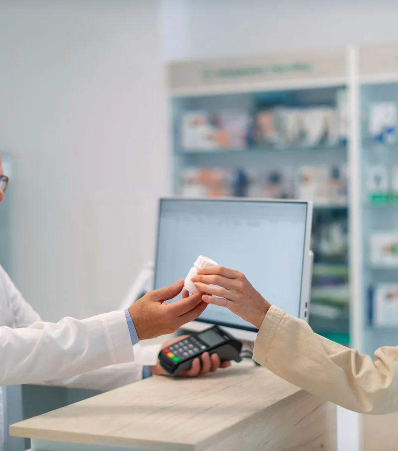 Pharmacist handing a prescription pill bottle to a customer at a pharmacy counter with a payment terminal and computer in the background.