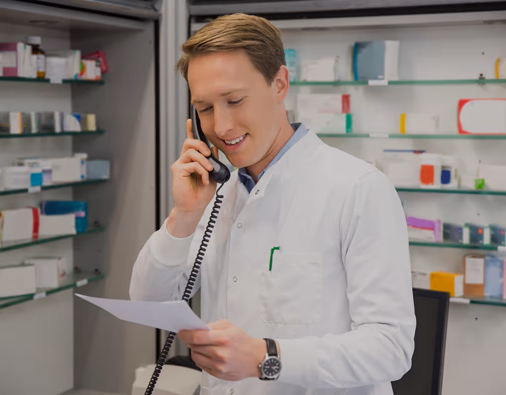 Pharmacist in a white coat talking on the phone and reading a paper inside a pharmacy.