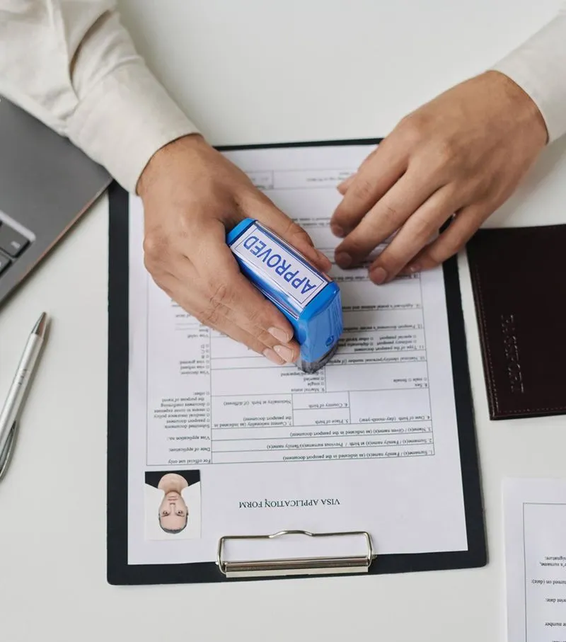 Person stamping an approved visa application form on a clipboard with a blue 'APPROVED' stamp.