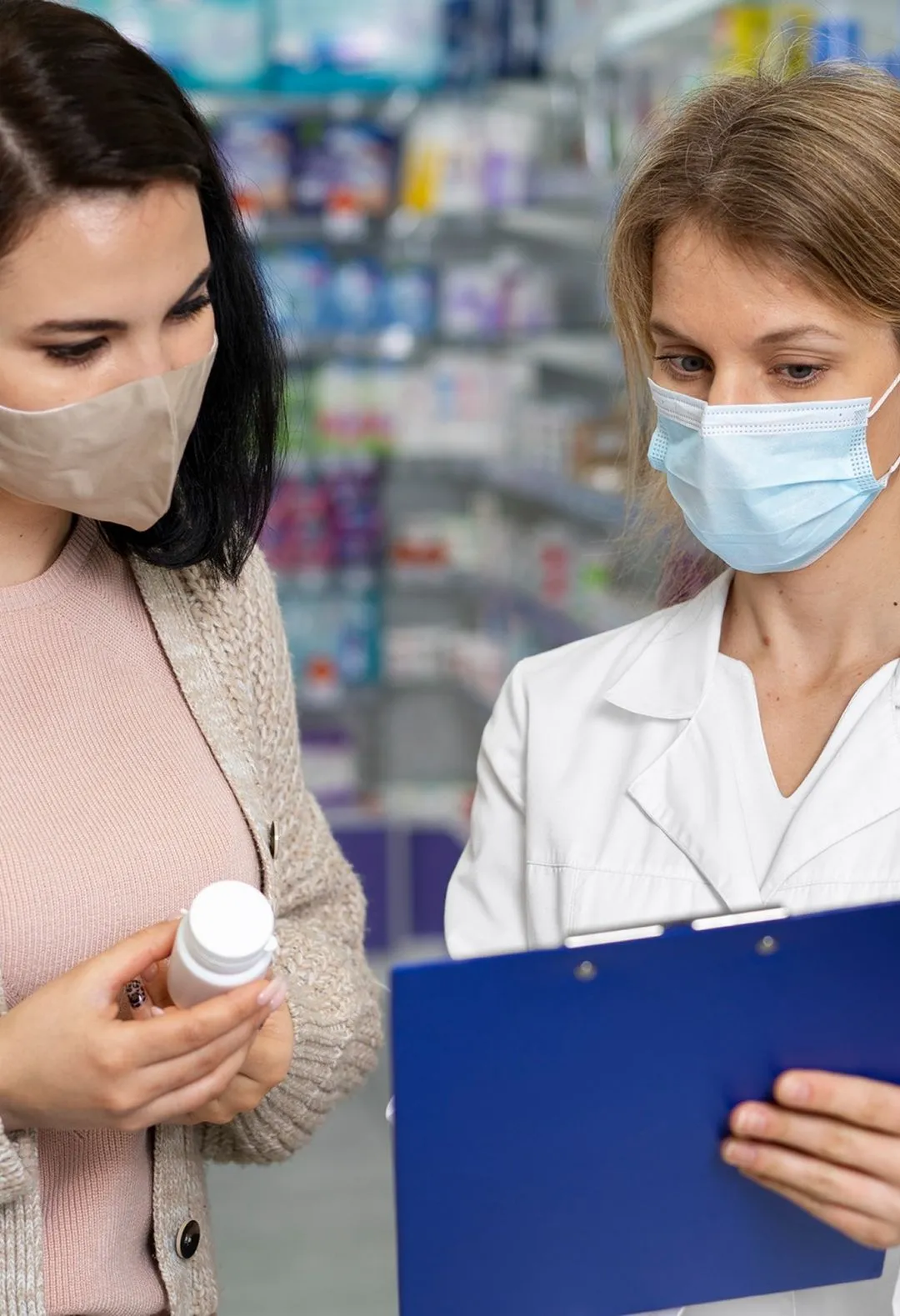 Pharmacist in white coat and mask consulting a masked customer holding a medicine bottle in a pharmacy.