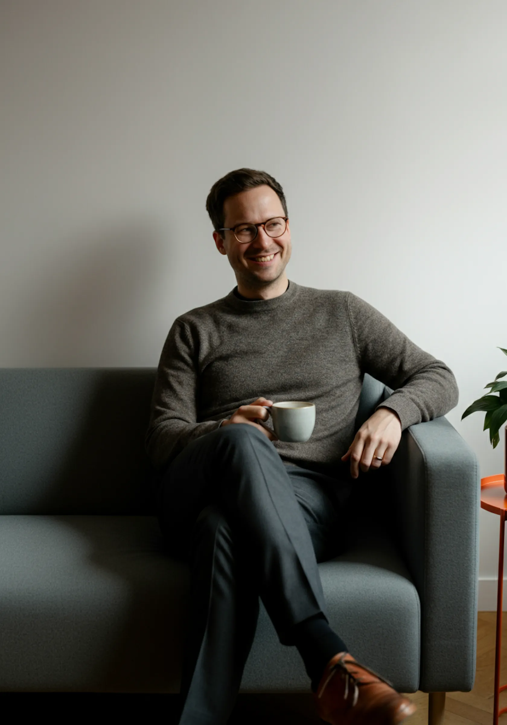 Man with glasses smiling while sitting cross-legged on a gray couch, holding a coffee cup.
