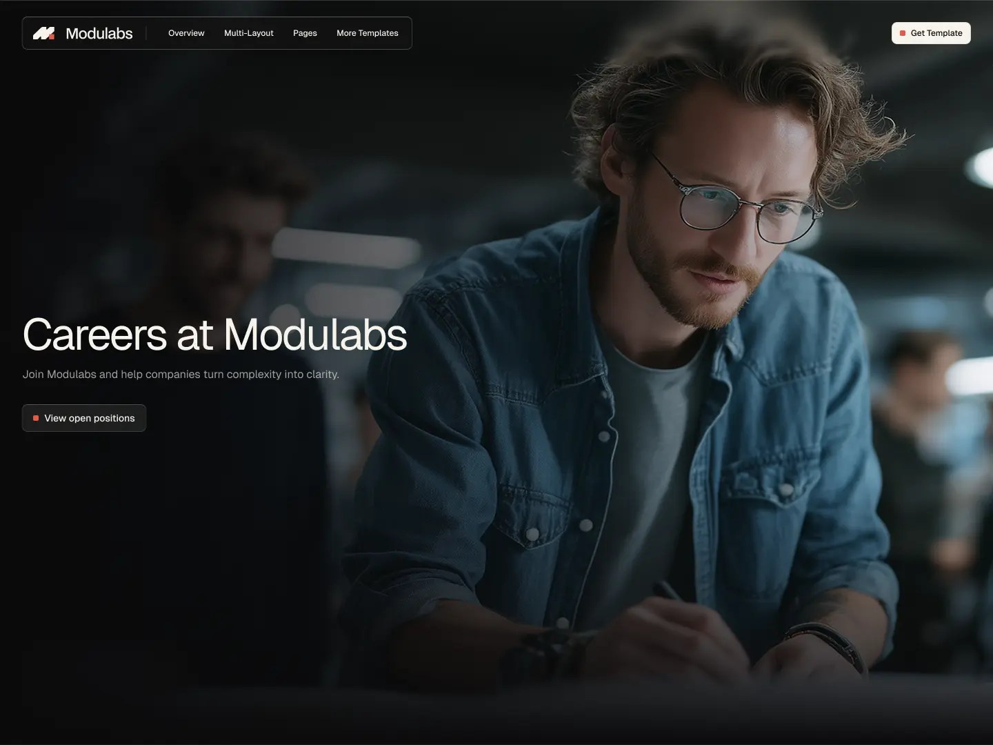 Man with glasses and curly hair focused on work at a desk in an office setting with blurred people in background.