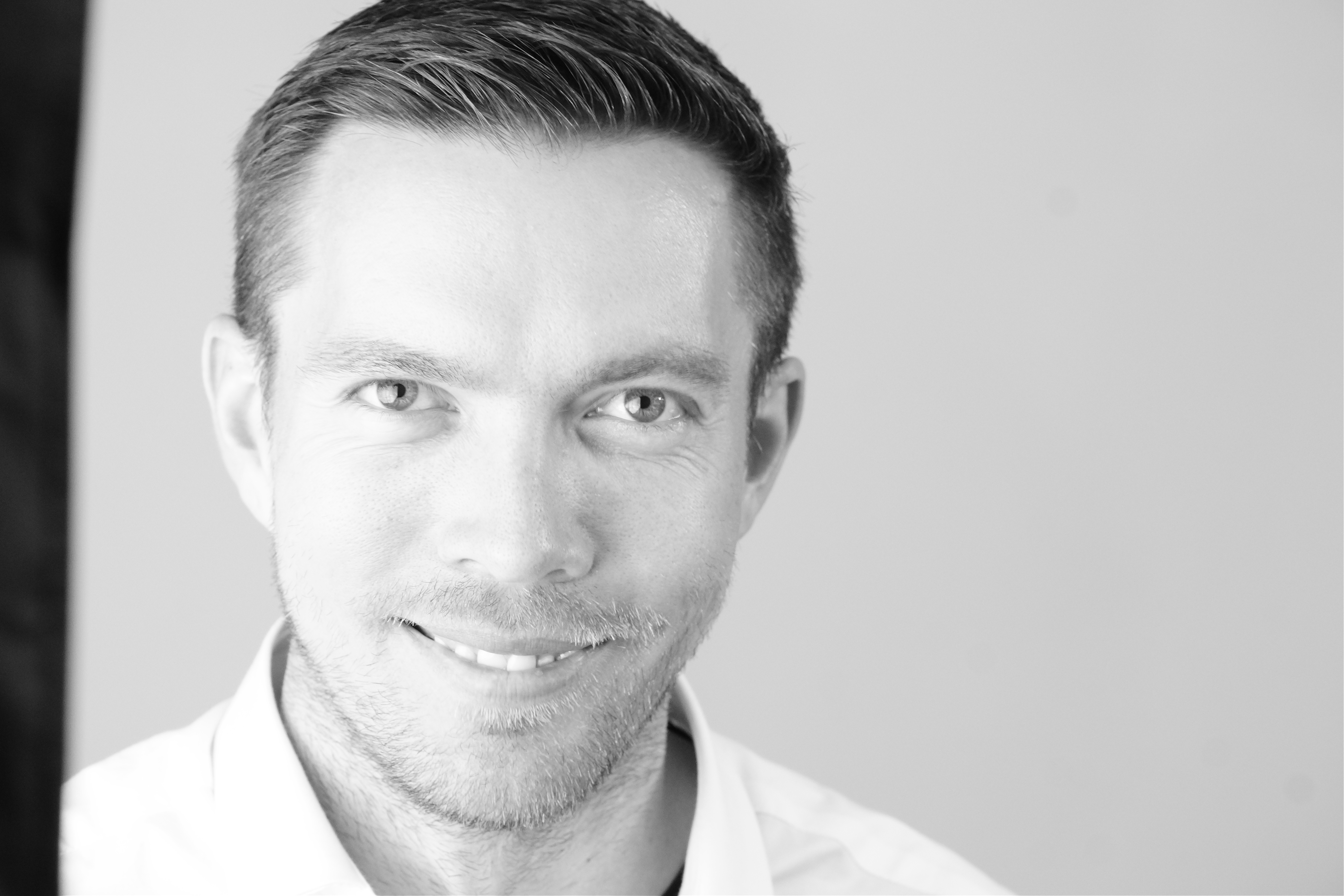 Black and white close-up portrait of a smiling man with short hair and light stubble.