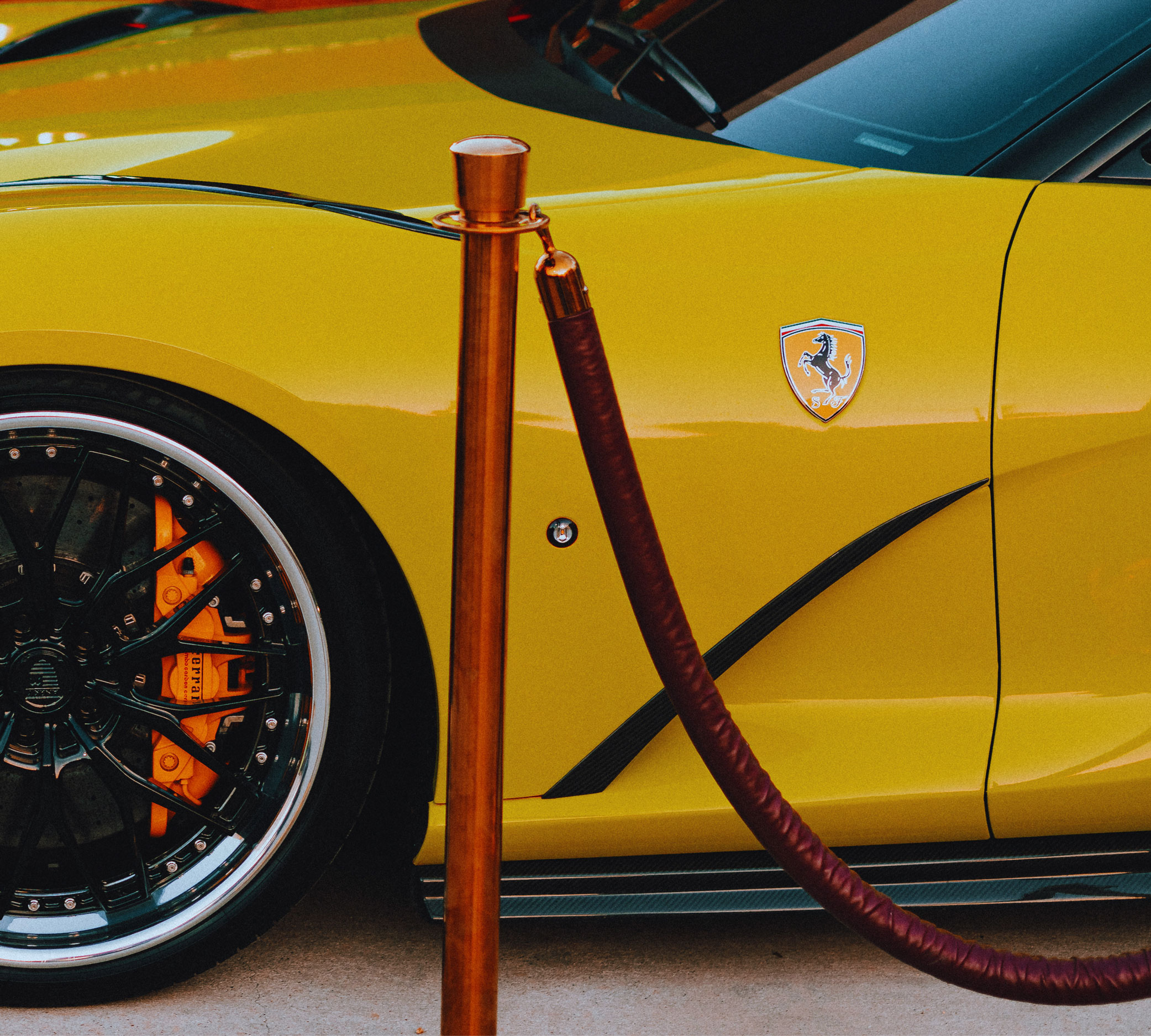Close-up of yellow Ferrari car with black wheel and orange brake caliper behind red velvet rope.