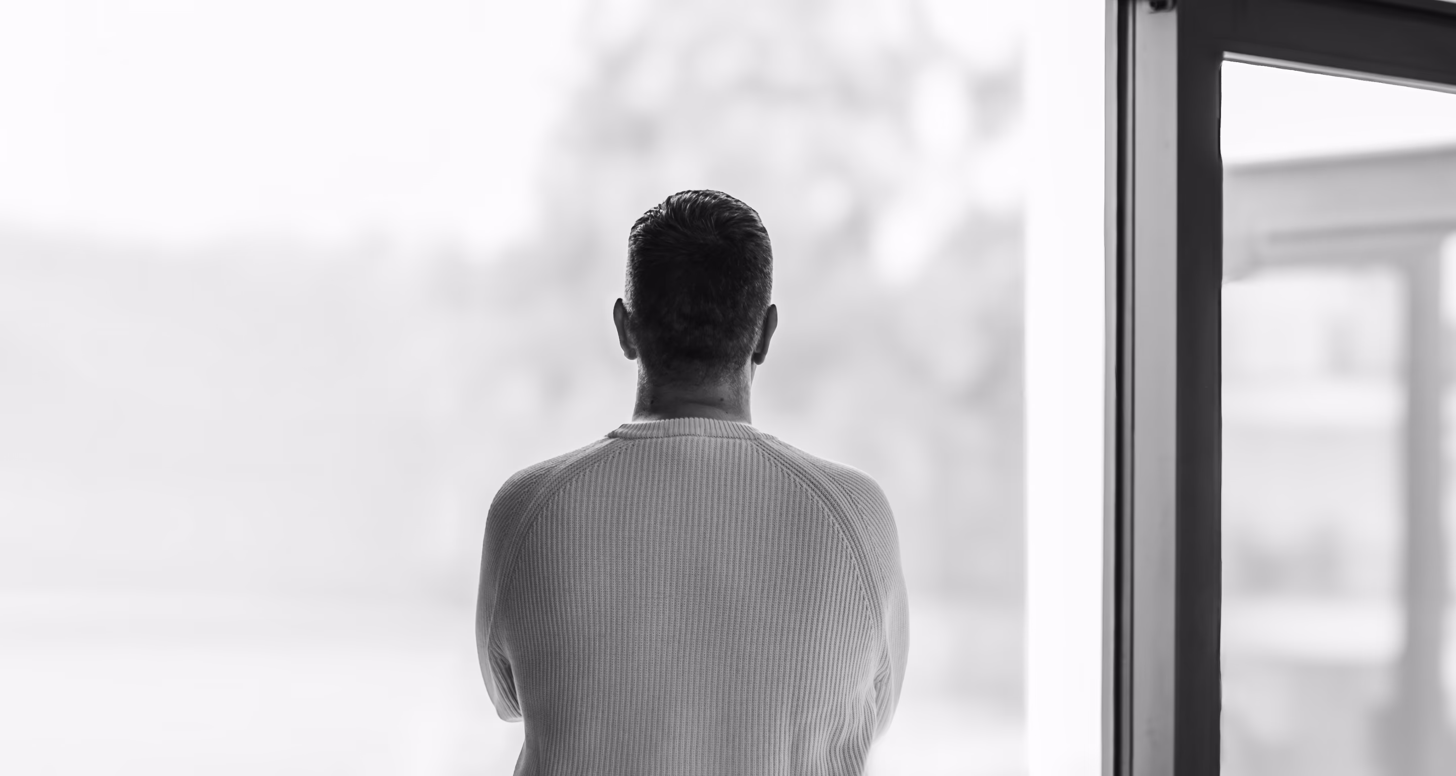 Black and white image of a man with short hair in a sweater looking out of a large window with a blurred outdoor background.