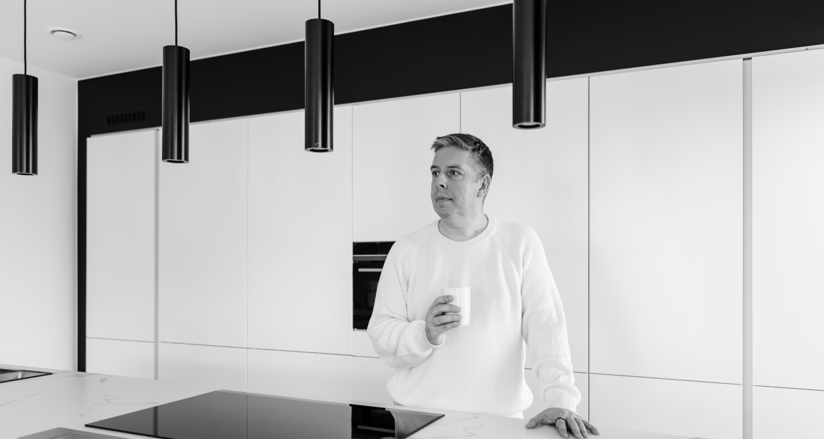 Man in a white sweater holding a cup in a modern kitchen with white cabinetry and black pendant lights.