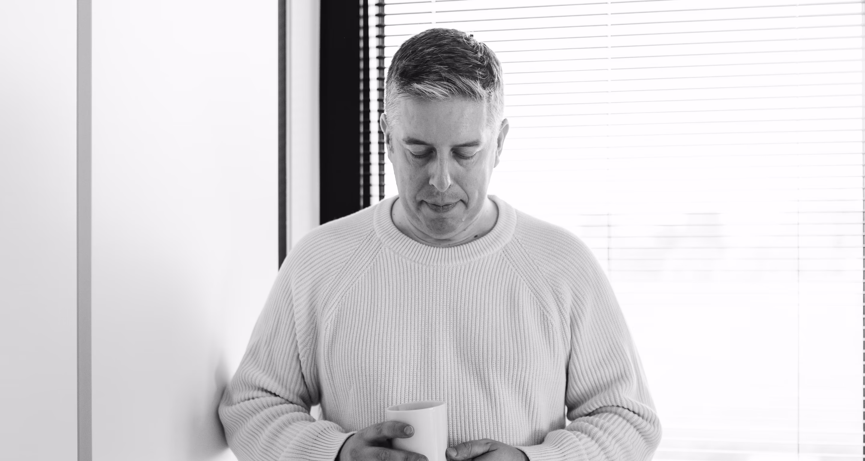 Middle-aged man wearing a textured sweater looking down while holding a cup with both hands near a window with blinds.