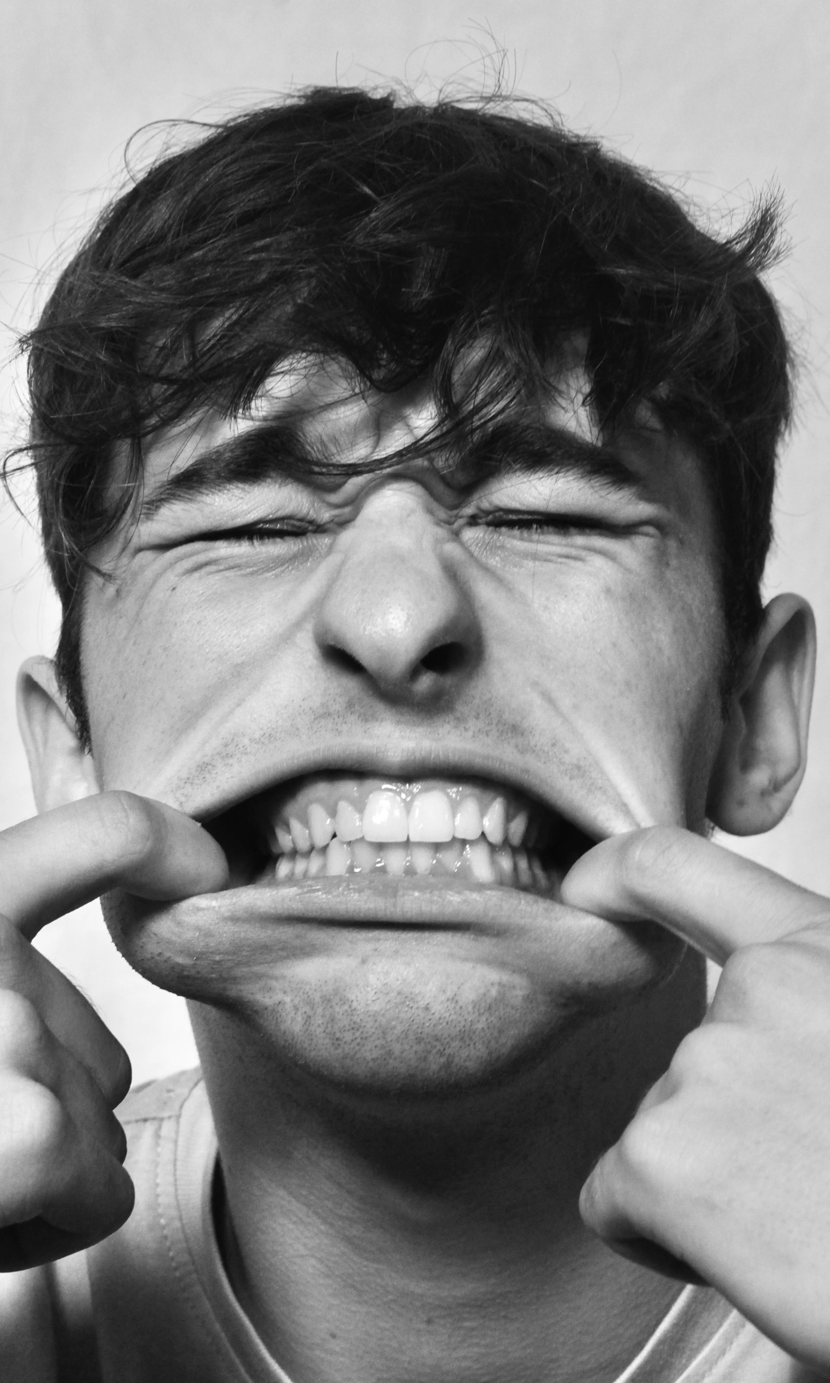 Close-up black and white photo of a man pulling his mouth wide to show his teeth with eyes closed tightly.