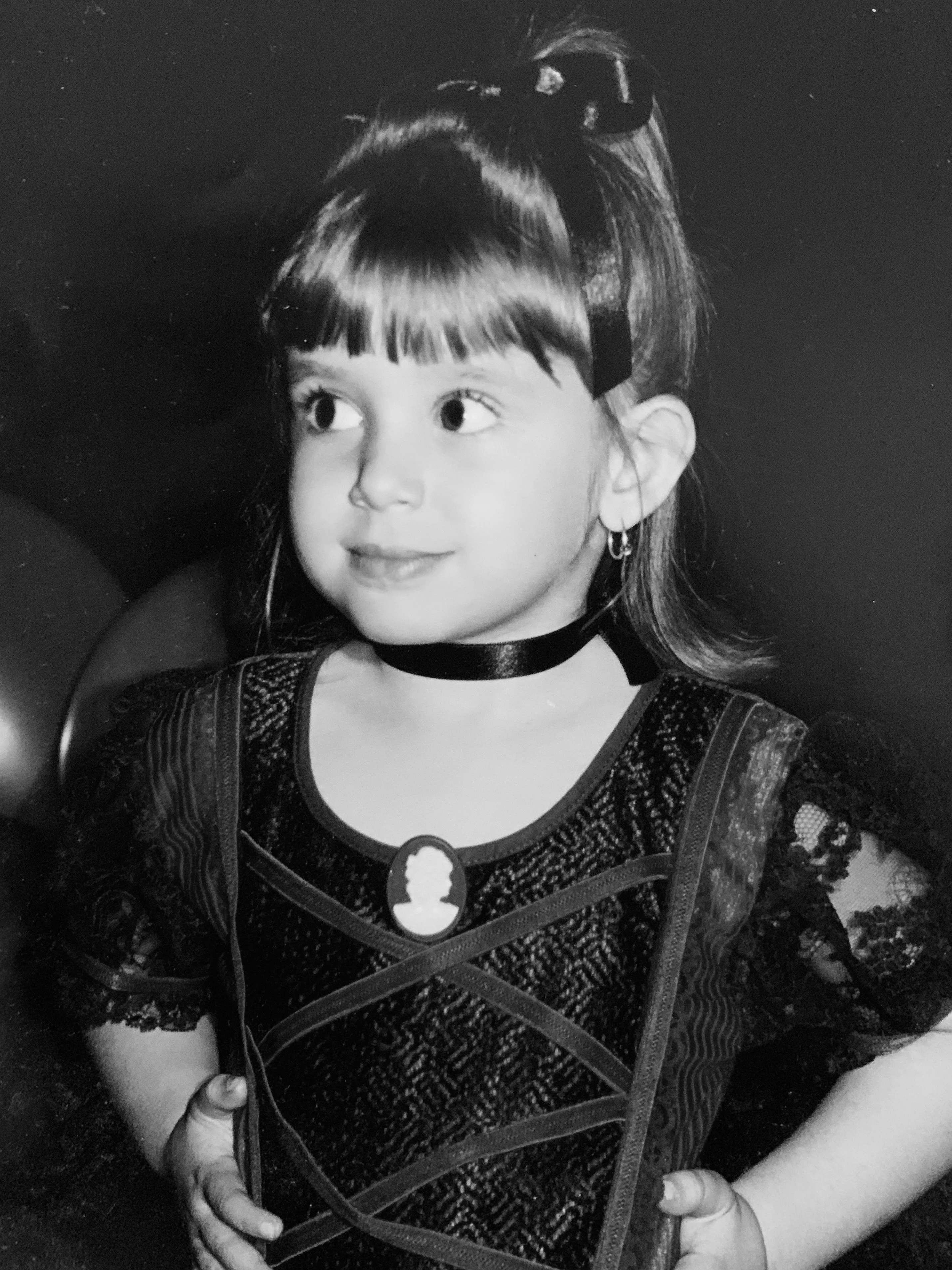 Black and white portrait of a young Rosanna with bangs and a ribbon in her hair, wearing a lace dress with a cameo brooch and choker necklace.