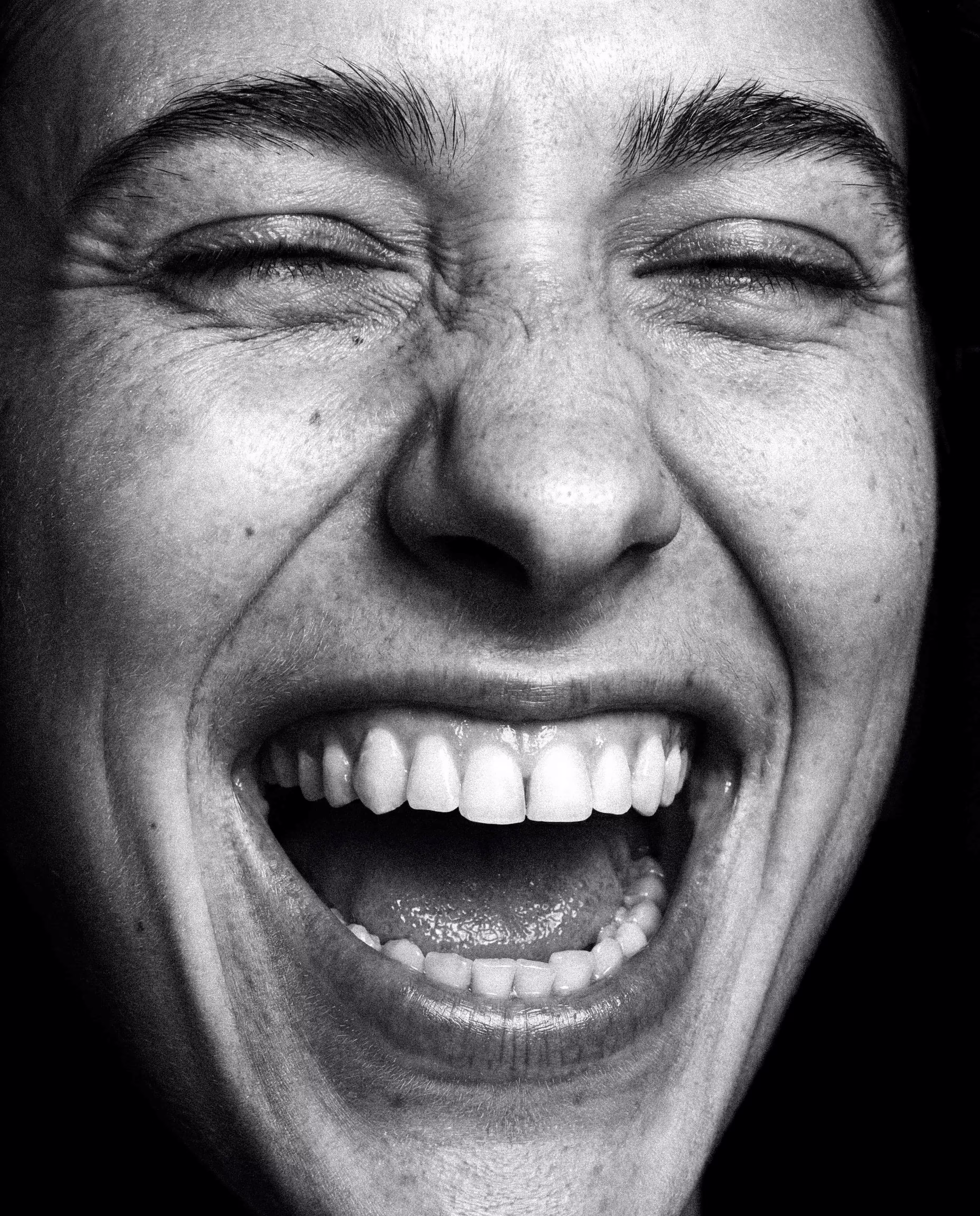Close-up black and white photo of a person with eyes closed and mouth wide open laughing.
