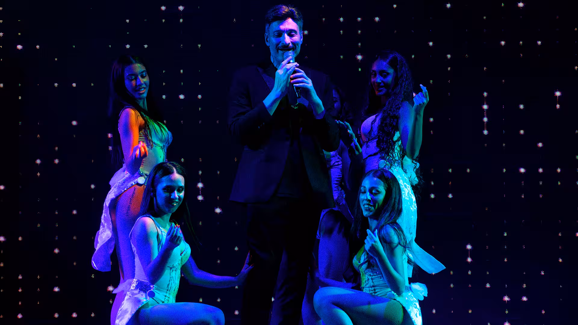 Male performer in black suit singing into a microphone surrounded by four female dancers in white outfits under blue stage lighting with a sparkling starry backdrop.