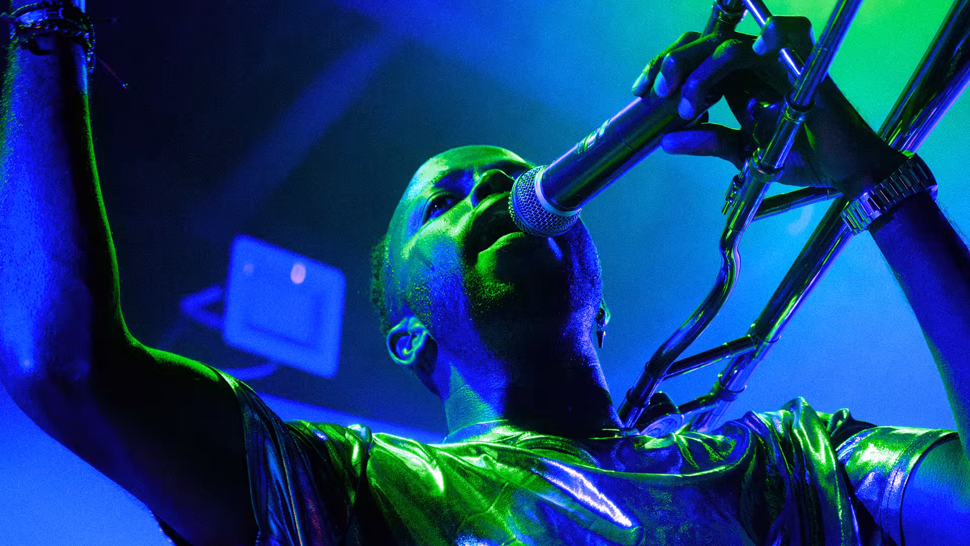 Musician singing into a microphone while playing a trombone under blue and green stage lighting.