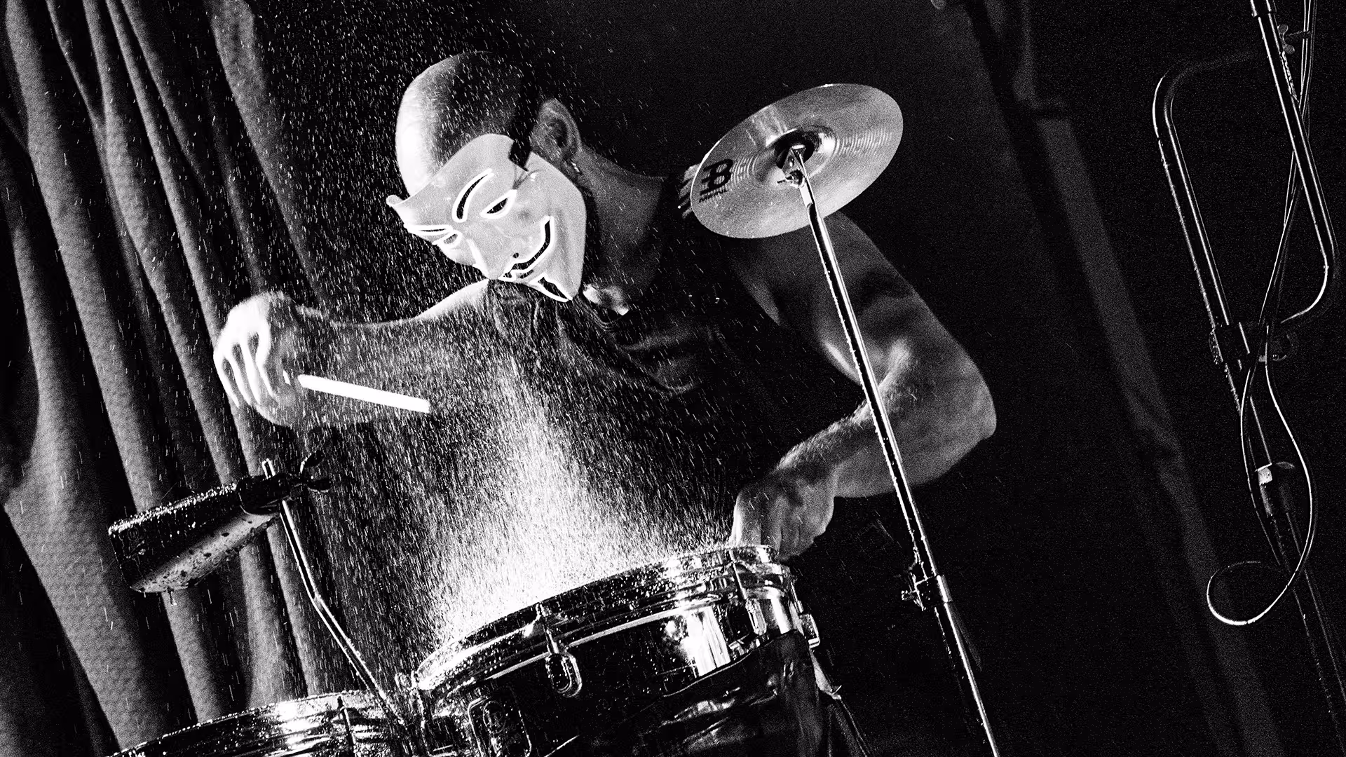 Black and white photo of a drummer wearing a Guy Fawkes mask playing drums with water splashing around.