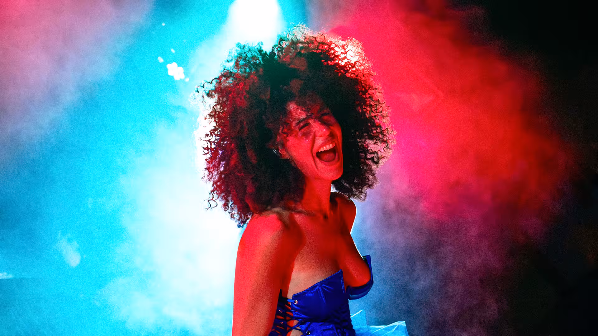 Woman with curly hair singing energetically under vibrant red and blue stage lights and smoke.
