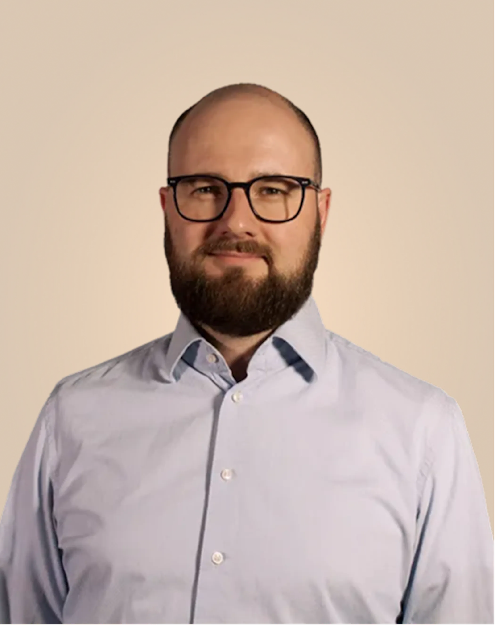 Portrait of a bald man with a beard and glasses wearing a light blue button-up shirt against a beige background.