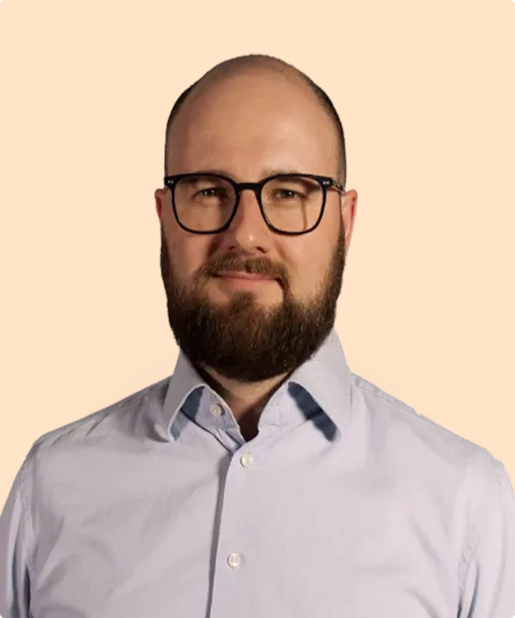 Bearded man with glasses wearing a light blue buttoned shirt against a beige background.