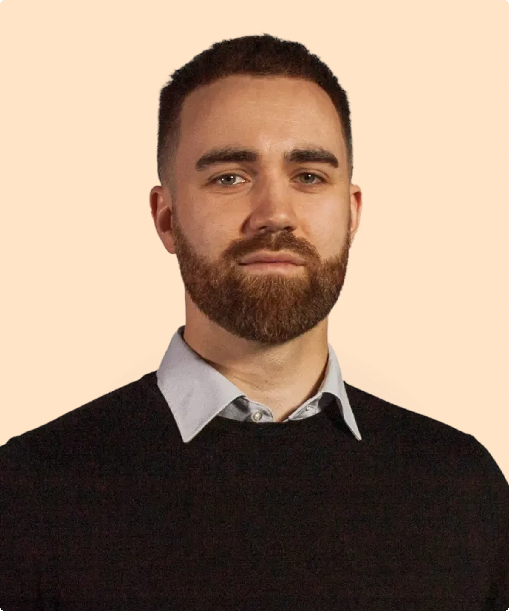 Portrait of a serious man with short hair, a full beard, wearing a light blue collared shirt under a black sweater.