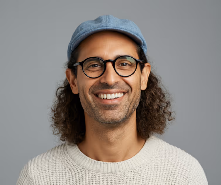 Smiling man with curly hair wearing round glasses, a light blue cap, and a white knitted sweater against a gray background.