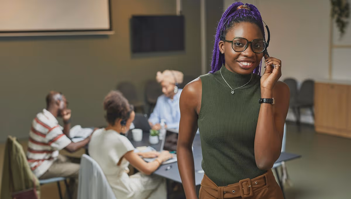 Smiling woman with purple braids and glasses wearing a headset in an office with three colleagues working in the background.