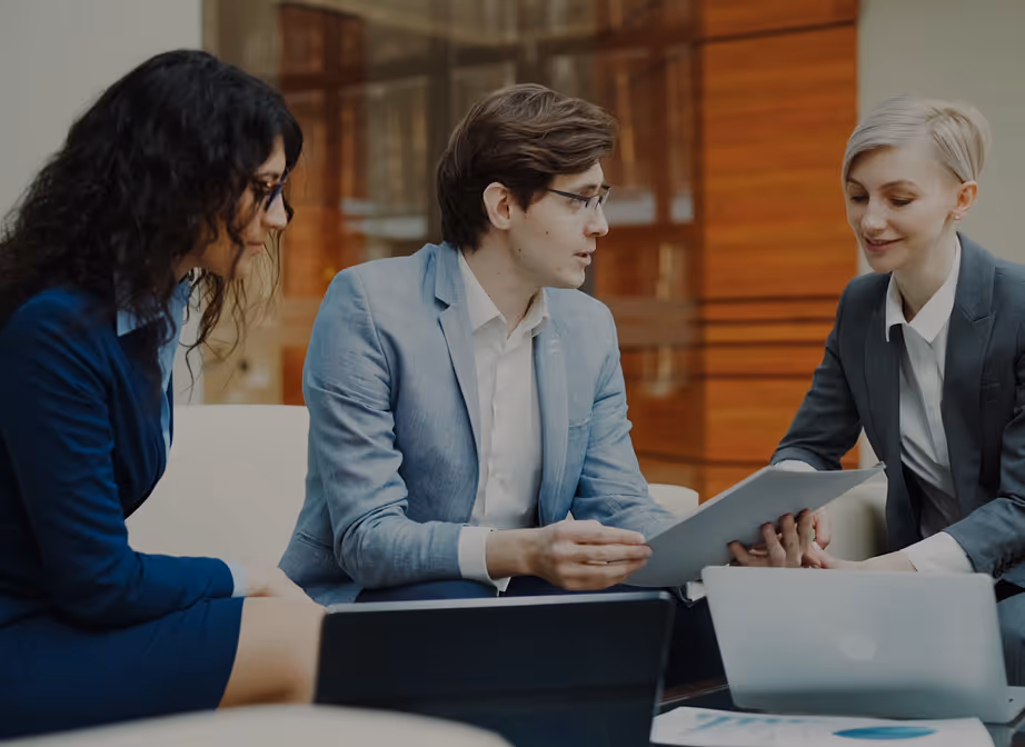 Three business professionals in formal attire sitting and discussing documents and laptops in a modern office setting.