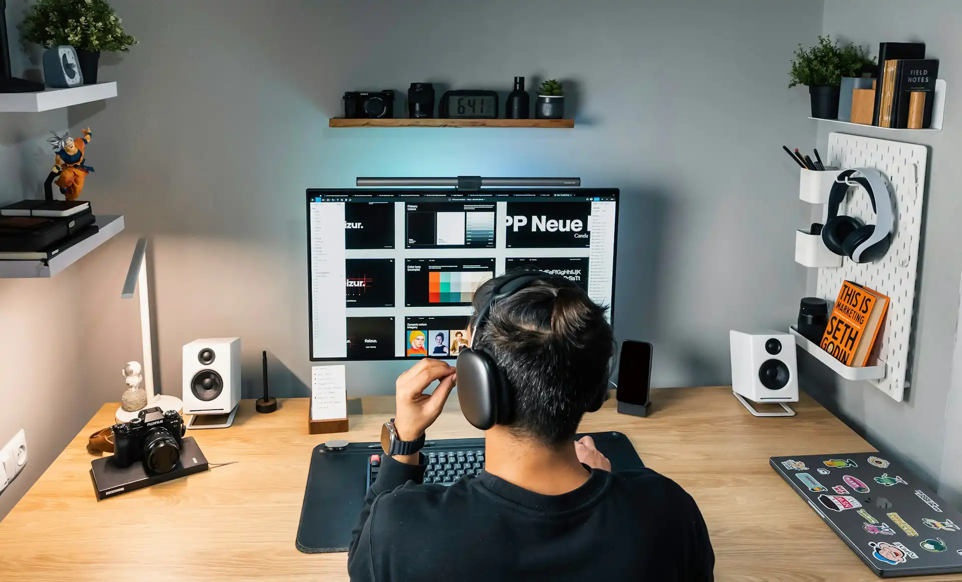 Person wearing headphones working at a desk with a large monitor, speakers, and a laptop with stickers.