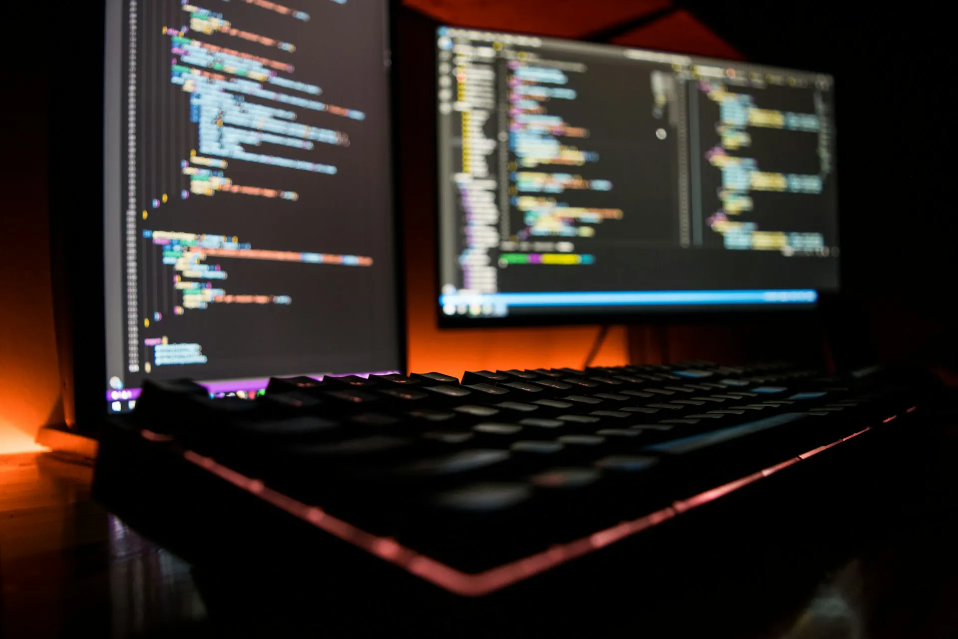 Close-up of a backlit keyboard with two monitors displaying blurred colorful programming code.