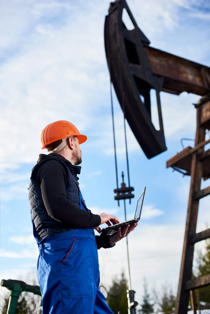 Worker in orange helmet and blue overalls operating laptop near an oil pumpjack under a cloudy sky.