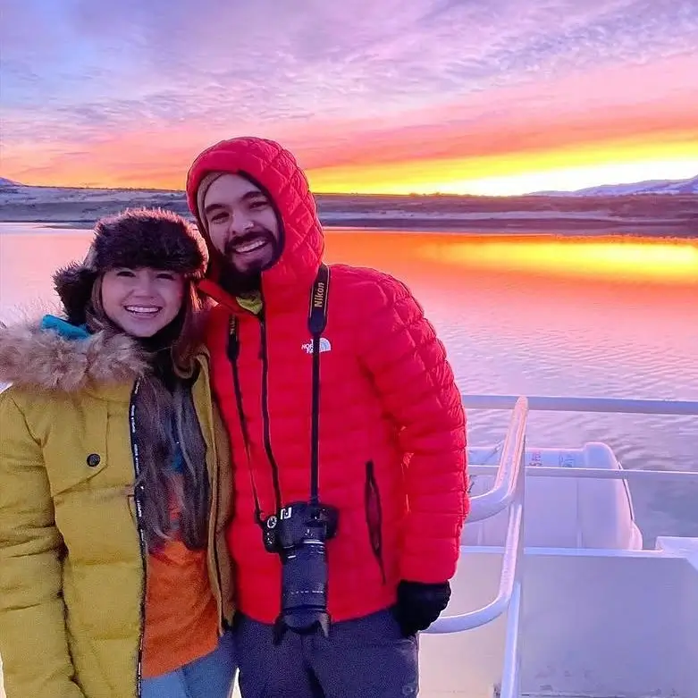 Milena y Carlos abrazados en un barco durante un atardecer ártico en tonos naranja y morado
