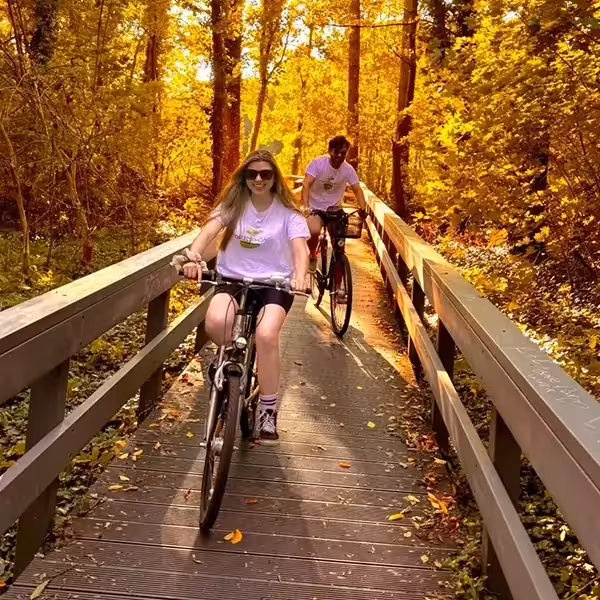 Milena y Carlos en bicicleta cruzando un puente de madera rodeado de árboles en otoño