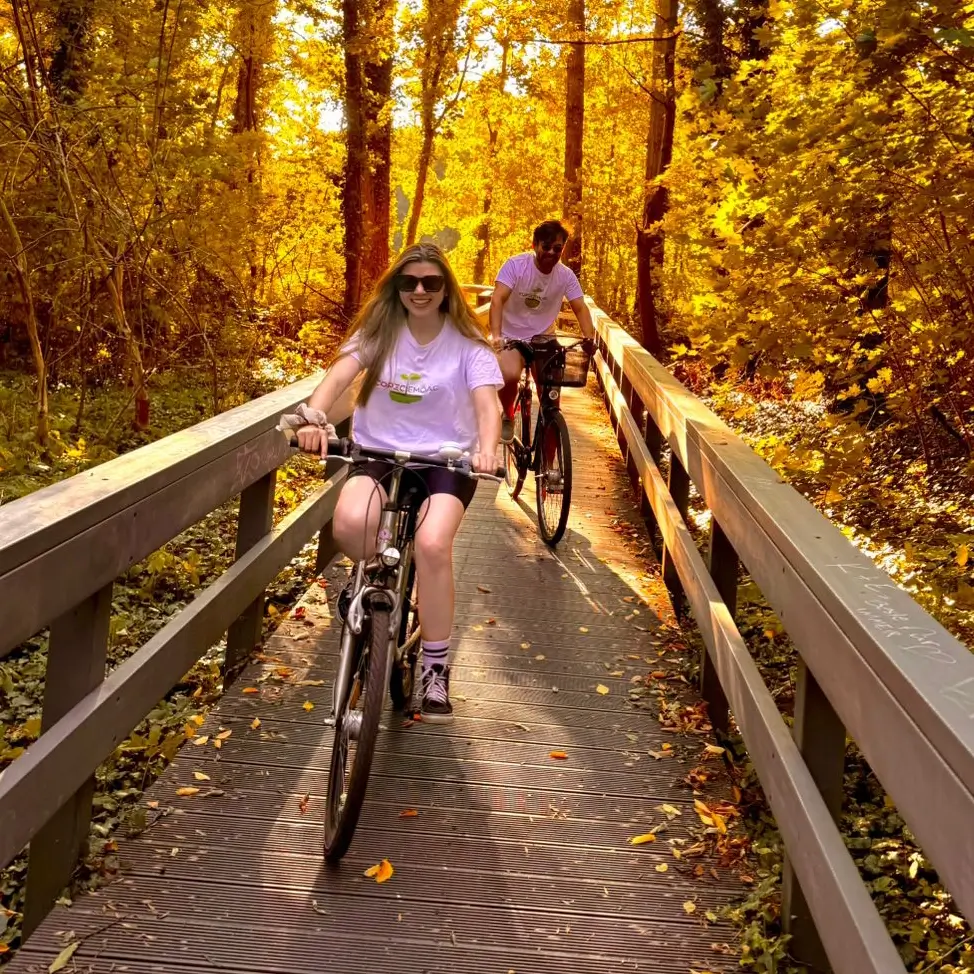 Milena y Carlos en bicicleta cruzando un puente de madera rodeado de árboles en otoño