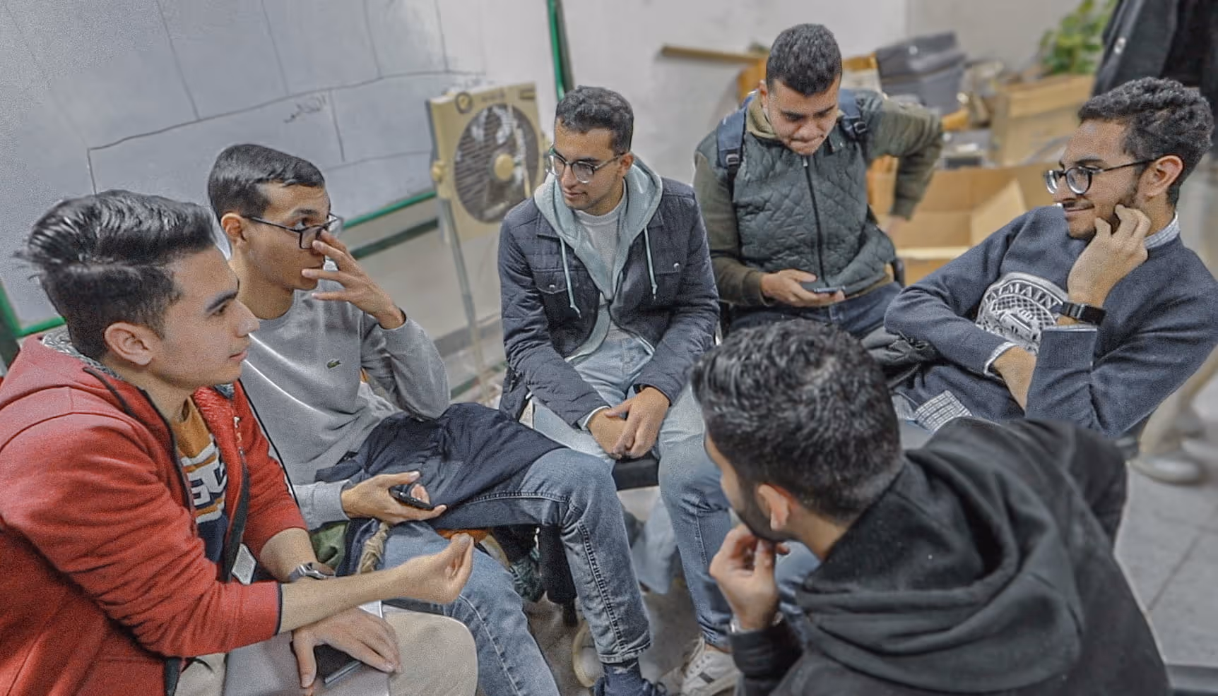 Group of six young men sitting indoors, engaged in conversation and using mobile phones.
