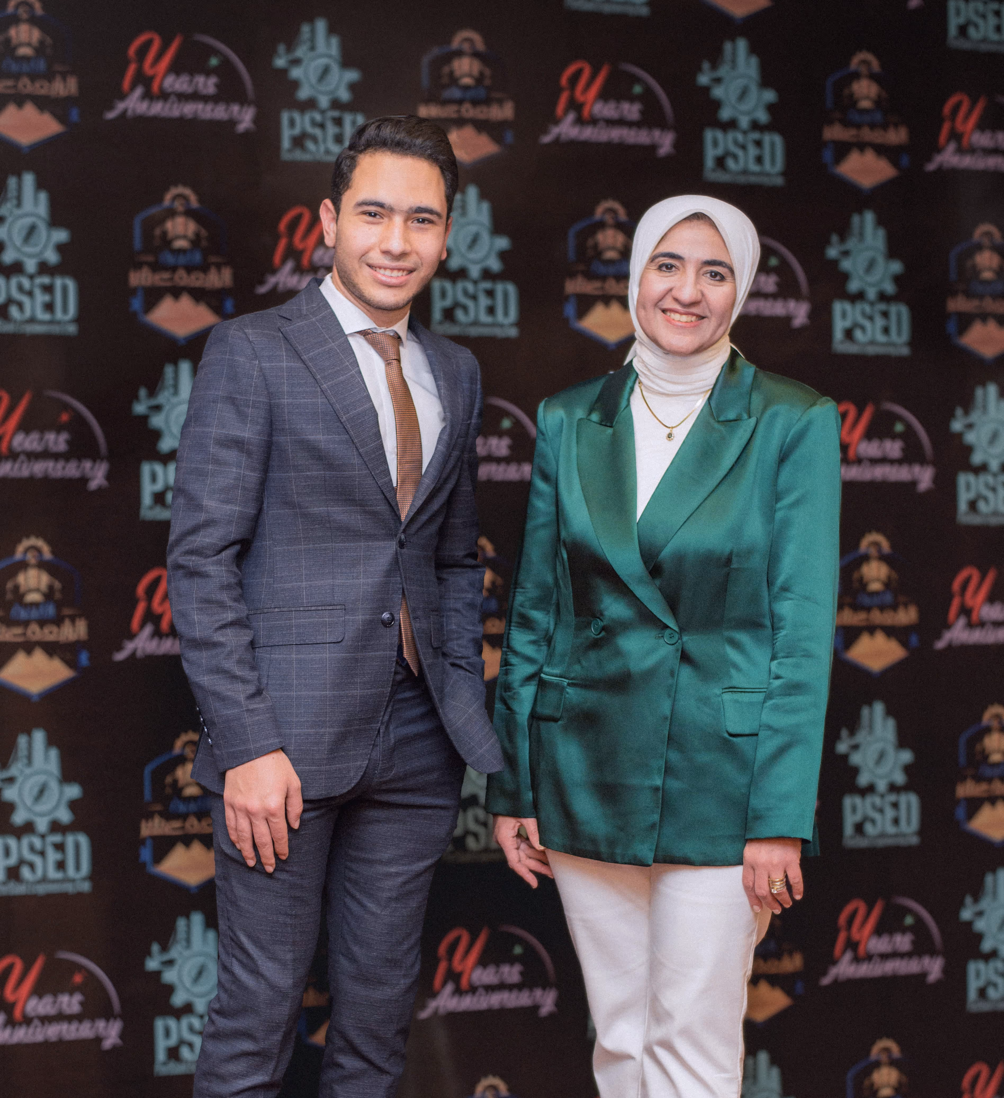 A man in a suit and a woman in a green blazer and white hijab smiling in front of an event backdrop.