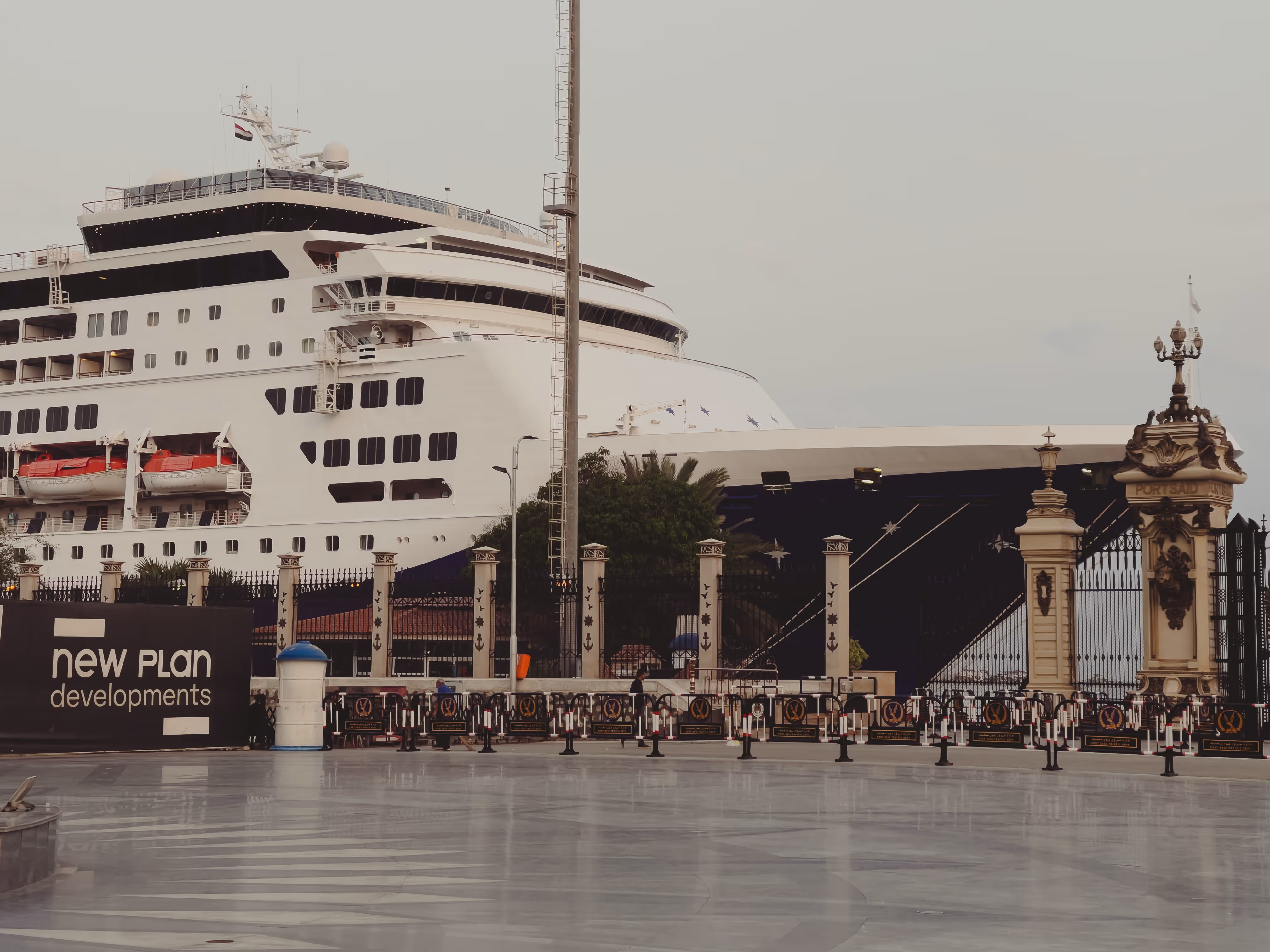 Large white cruise ship docked behind decorative iron fence with pillars and gate at a port.