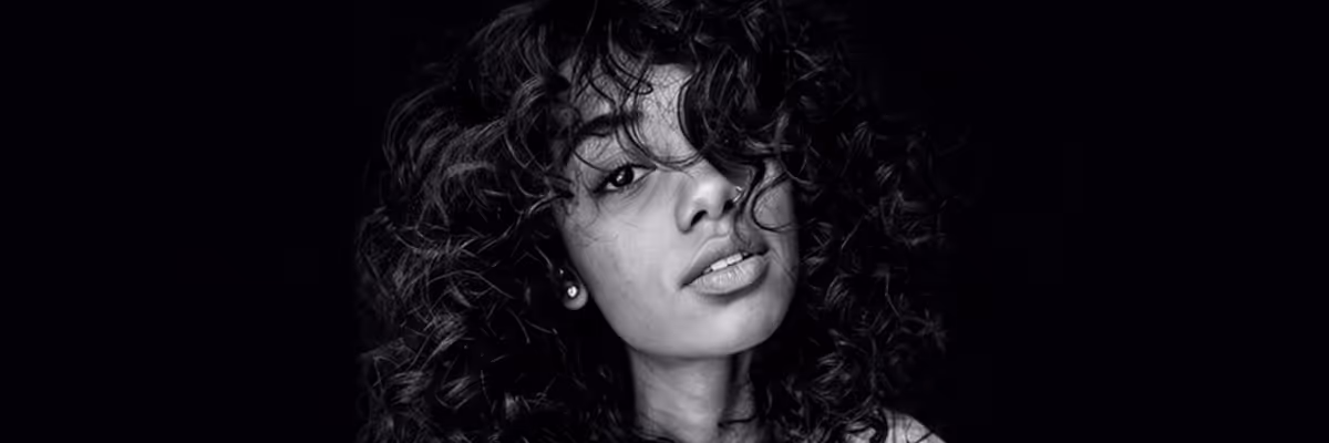 Black and white close-up of a woman with curly hair partially covering her face against a black background.