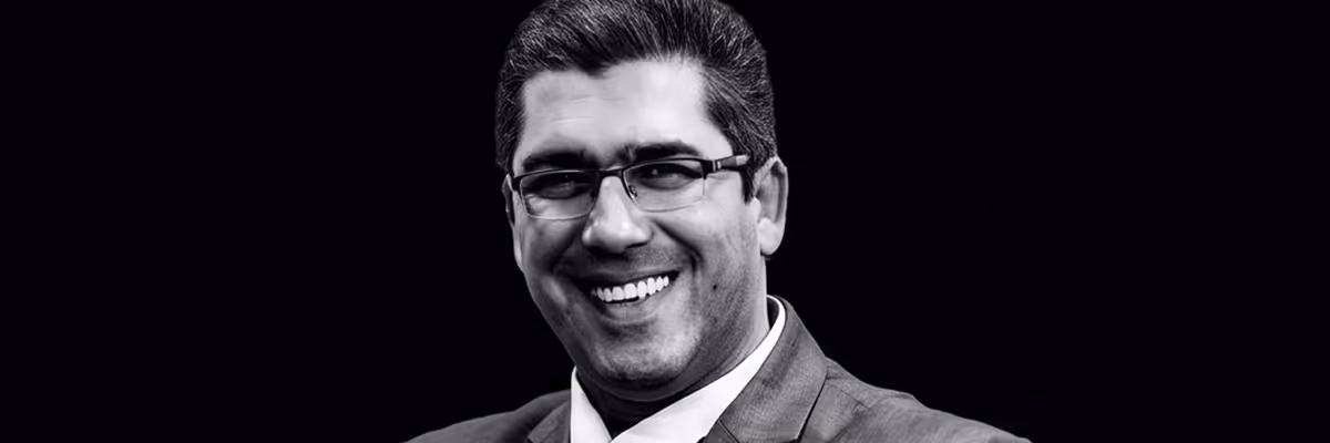 Smiling man wearing glasses and a suit with a white shirt on a black background.