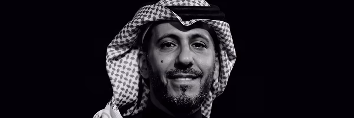 Black and white close-up portrait of a man wearing a traditional patterned headscarf with a slight smile.