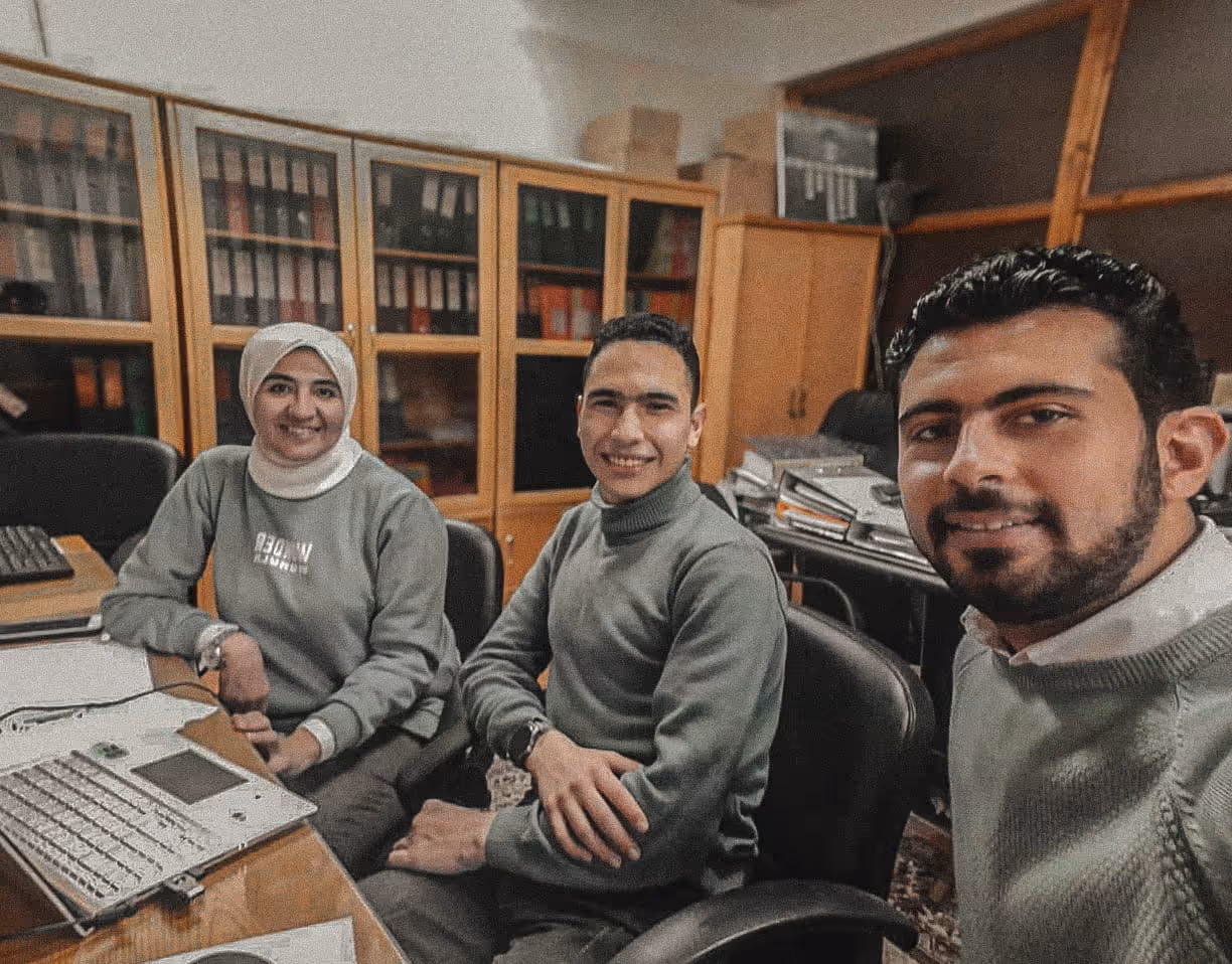 Three people sitting in an office smiling at the camera with a laptop and files on the desk.