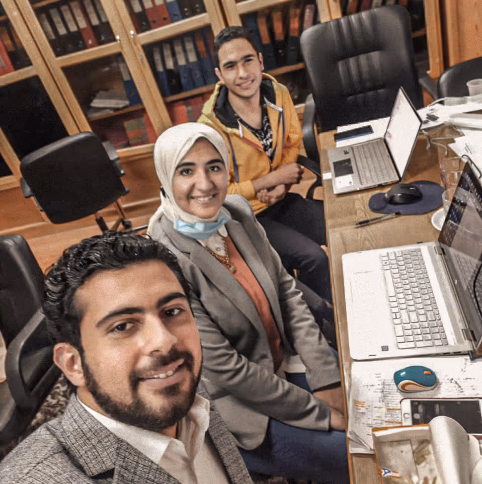 Three people smiling at camera in an office with laptops, papers, and bookshelves behind them.
