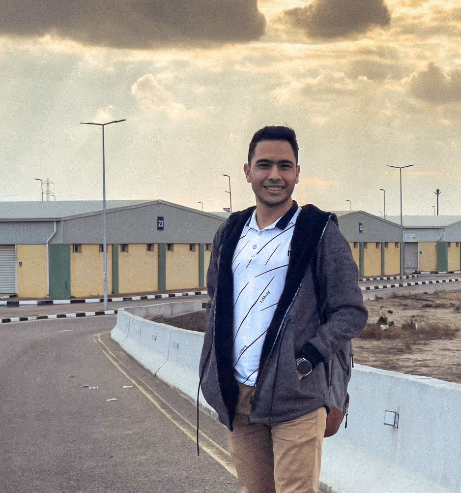 Smiling man in a gray jacket and backpack standing on a road near industrial buildings at sunset.
