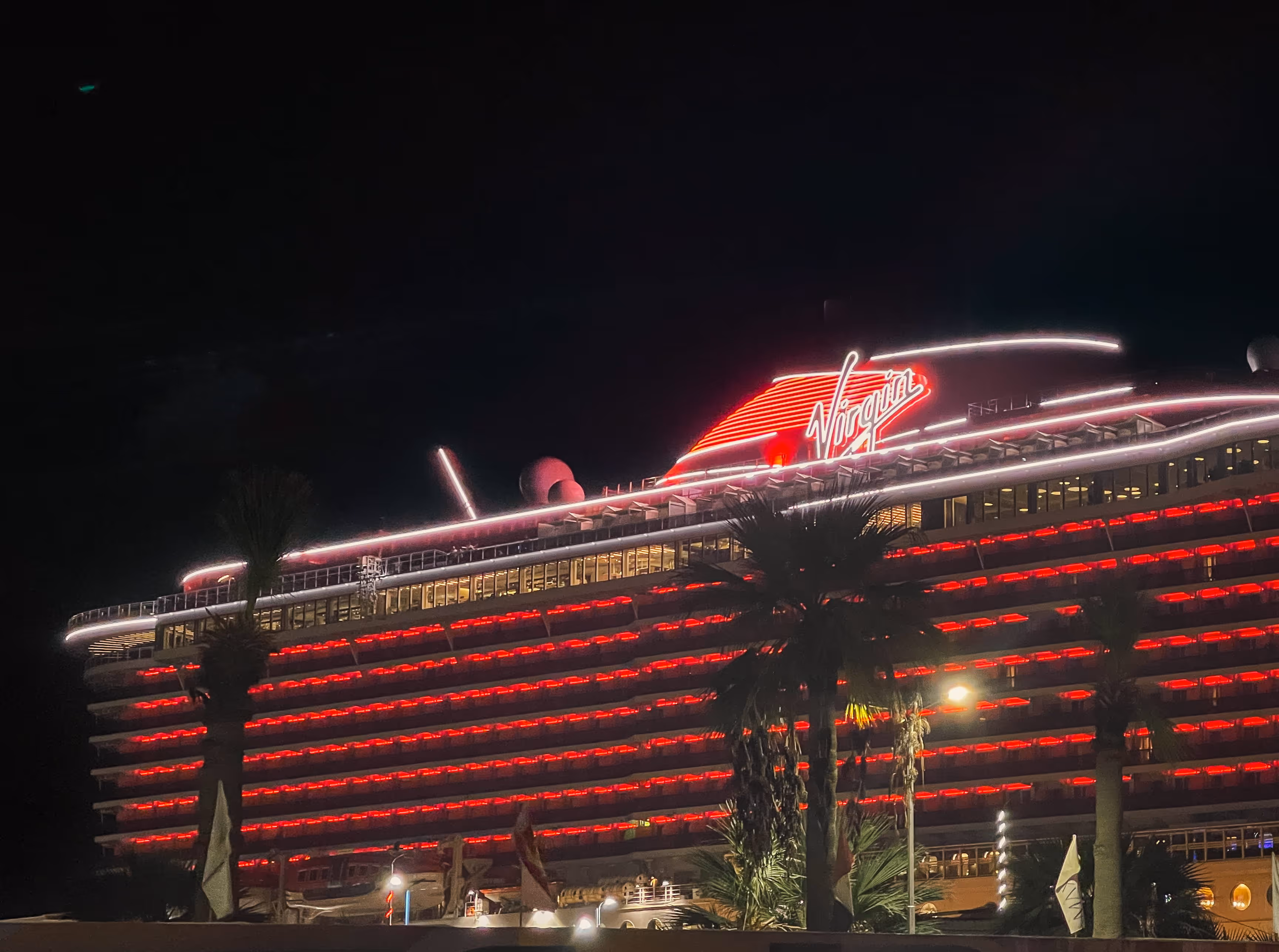 Virgin cruise ship illuminated at night with red and white lights, palm trees in foreground.