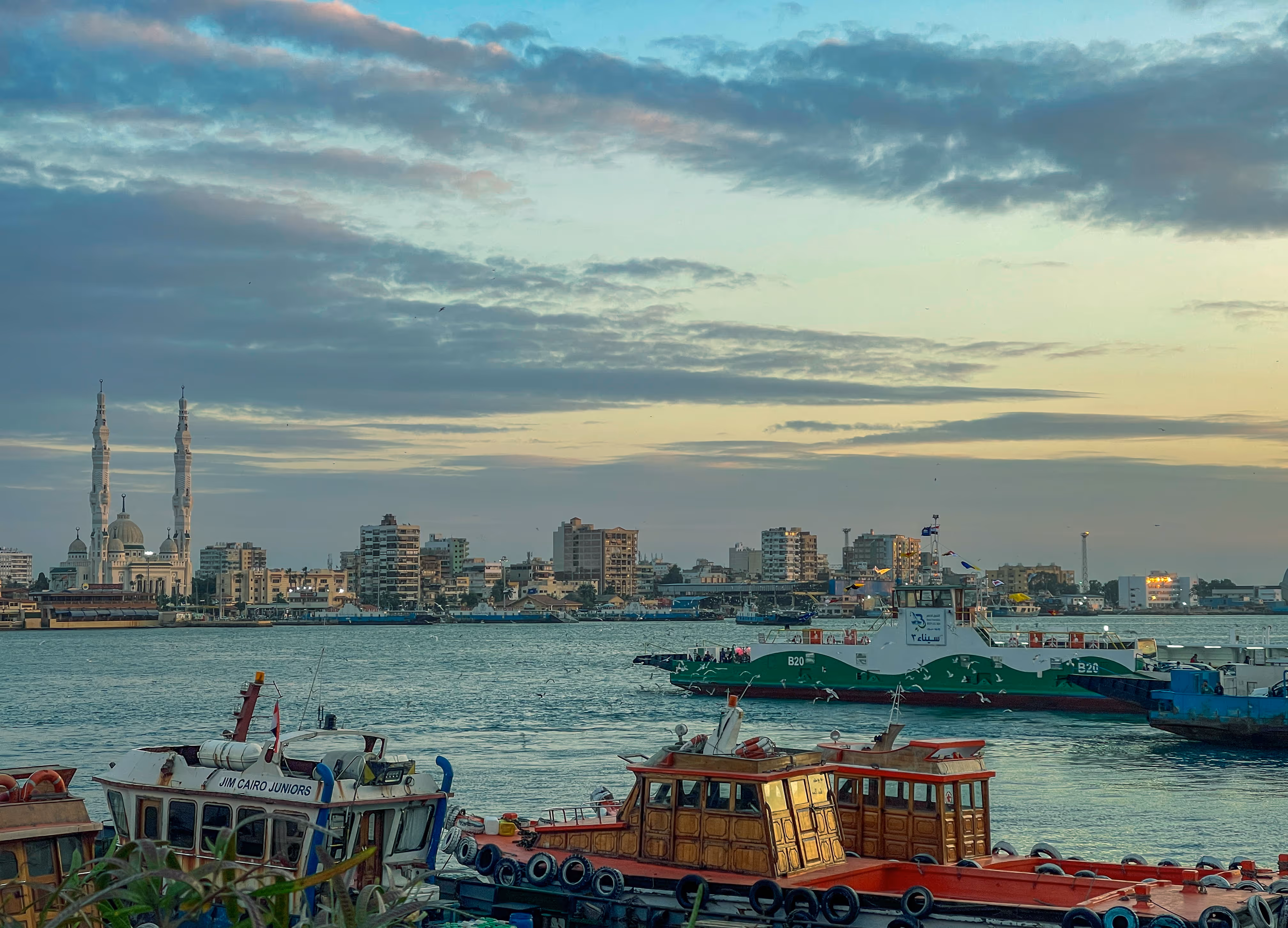 Port Said boats docked on a calm river with city buildings and a mosque in the background at sunset.