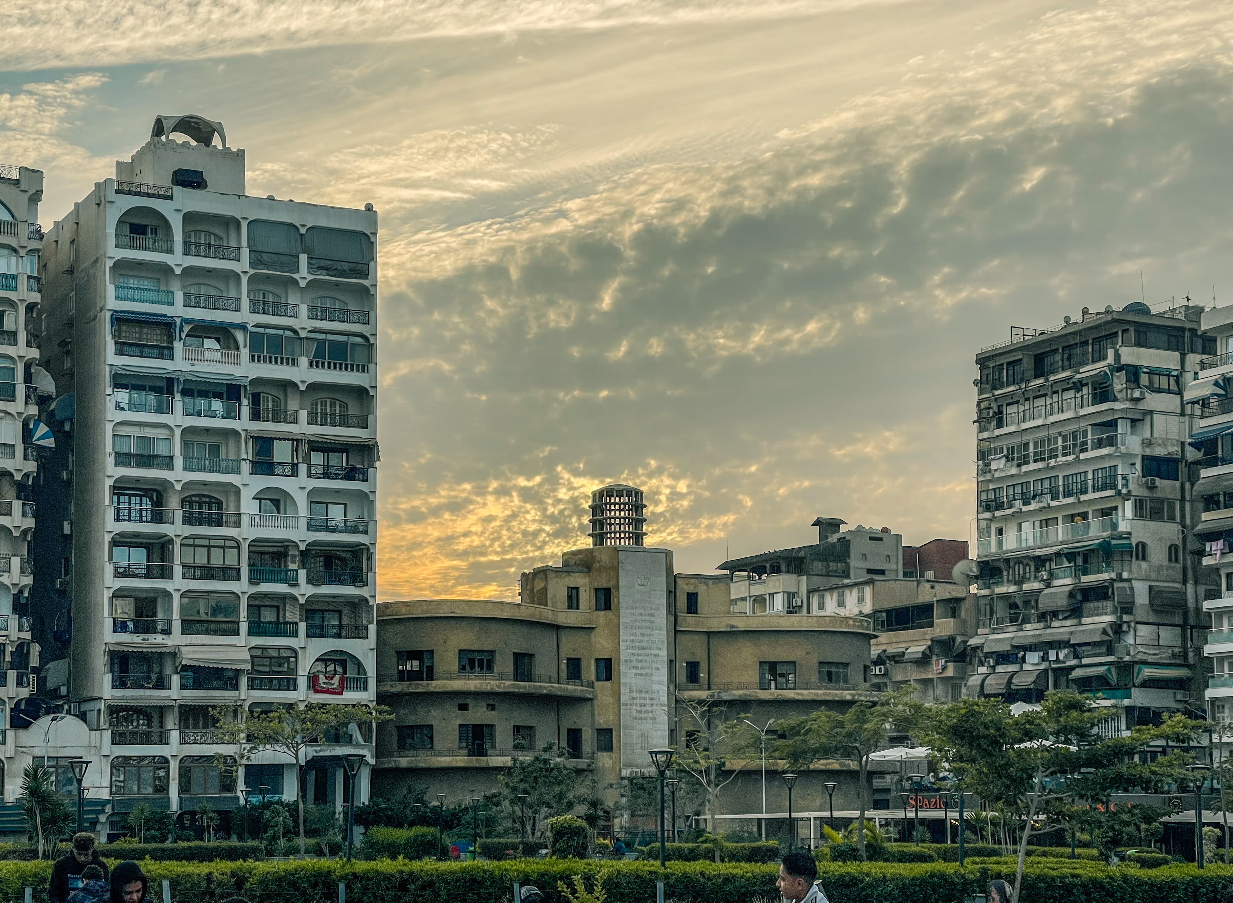 Urban buildings at sunset with cloudy sky and trees in the foreground.