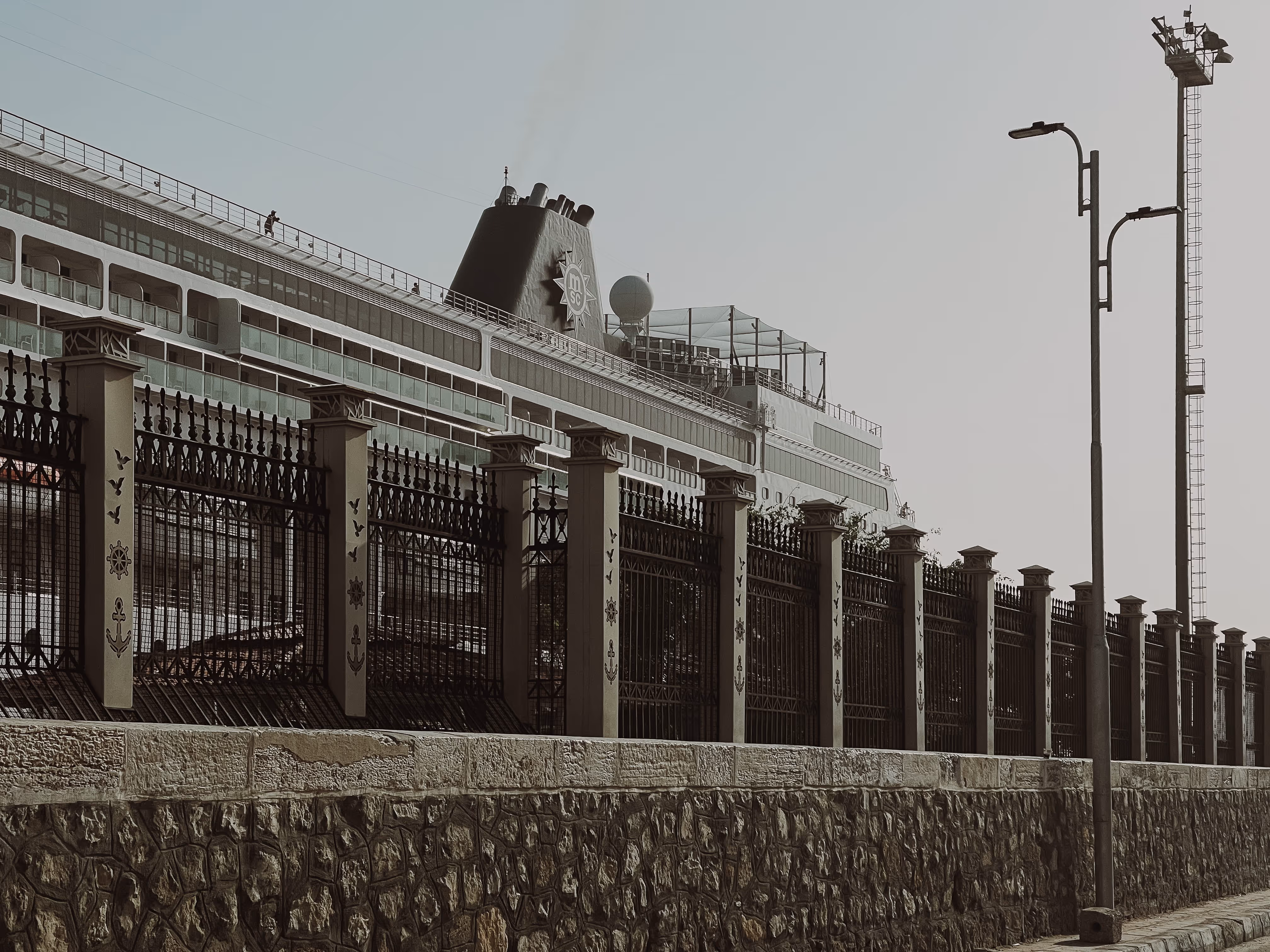 Large cruise ship docked behind a tall iron fence with stone base and street lamps nearby.