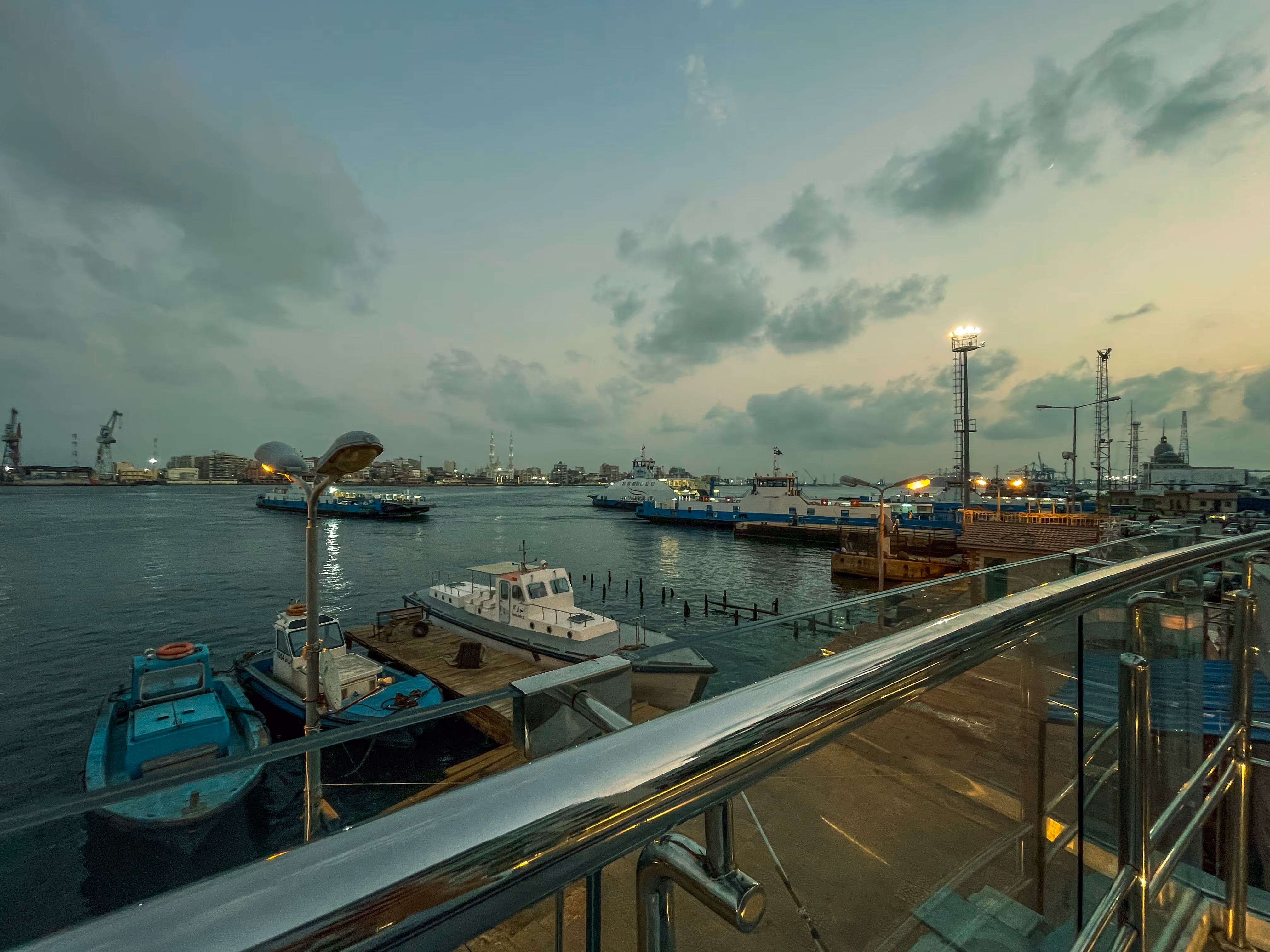 Harbor at dusk in Port Said with moored boats, lit lamp posts, industrial buildings, and cloudy sky.