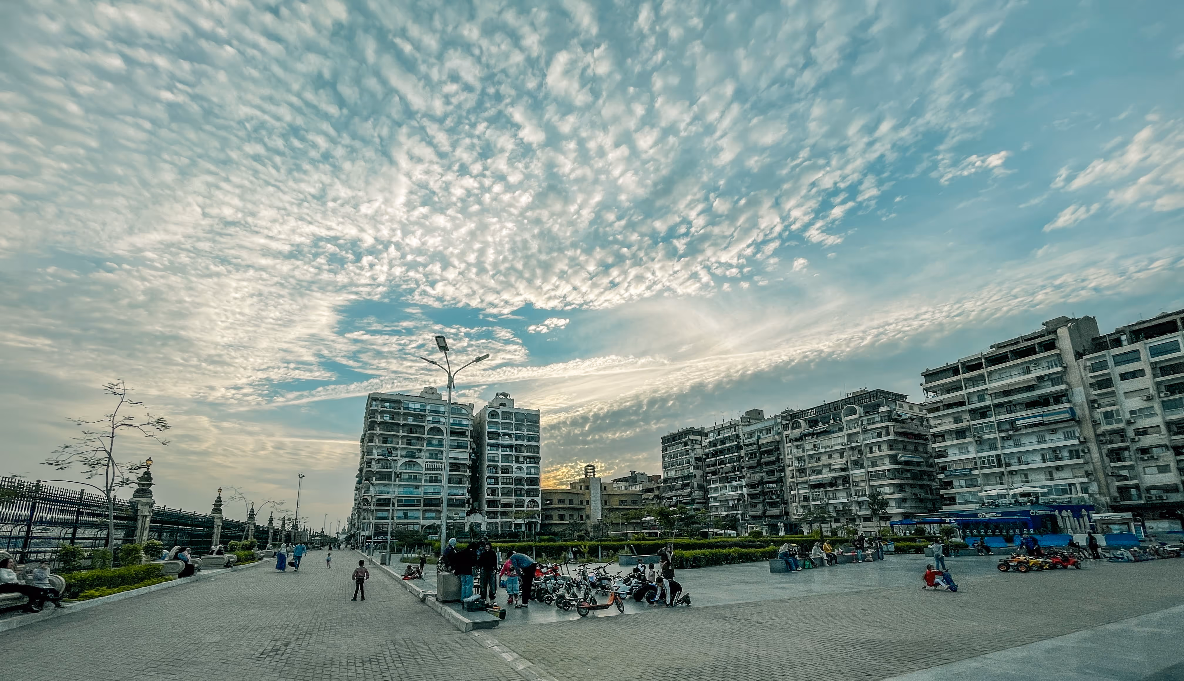 Urban plaza with people, motorcycles, and tall apartment buildings under a partly cloudy sky at sunset.