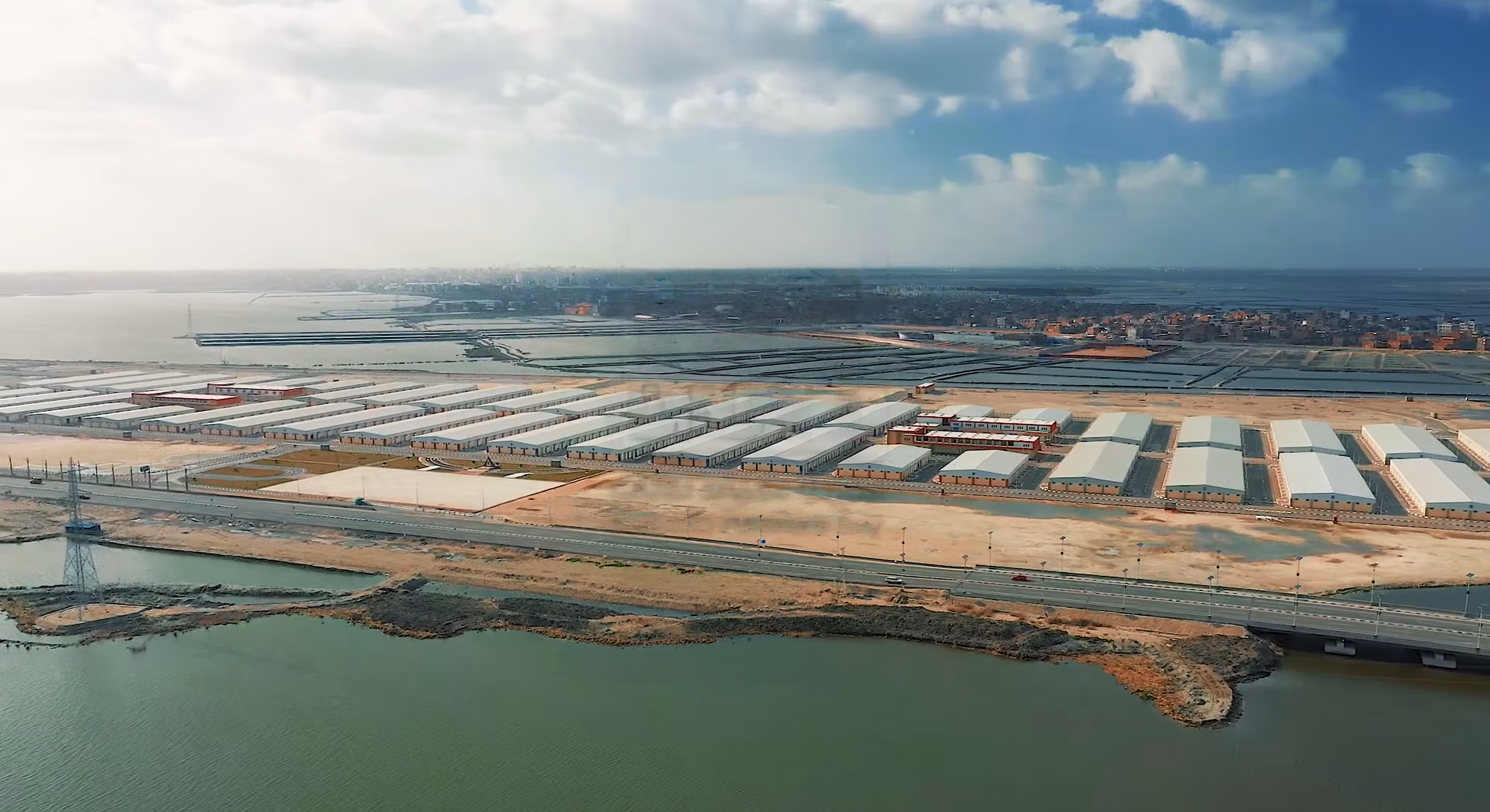 Aerial view of warehouses near water with a road and bridge in the foreground under a cloudy sky.
