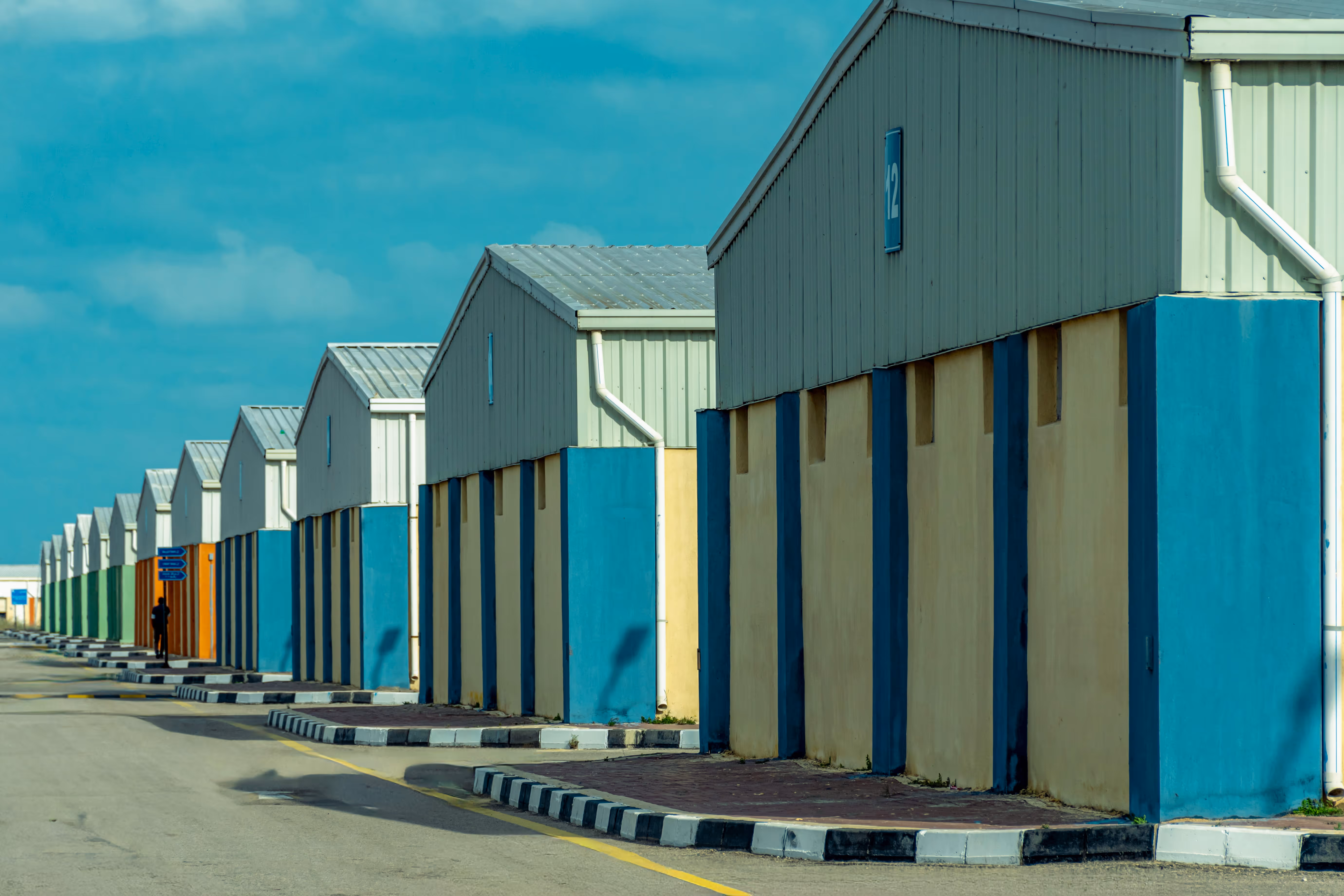 Row of warehouse buildings with blue, green, and orange sections under a daytime blue sky.